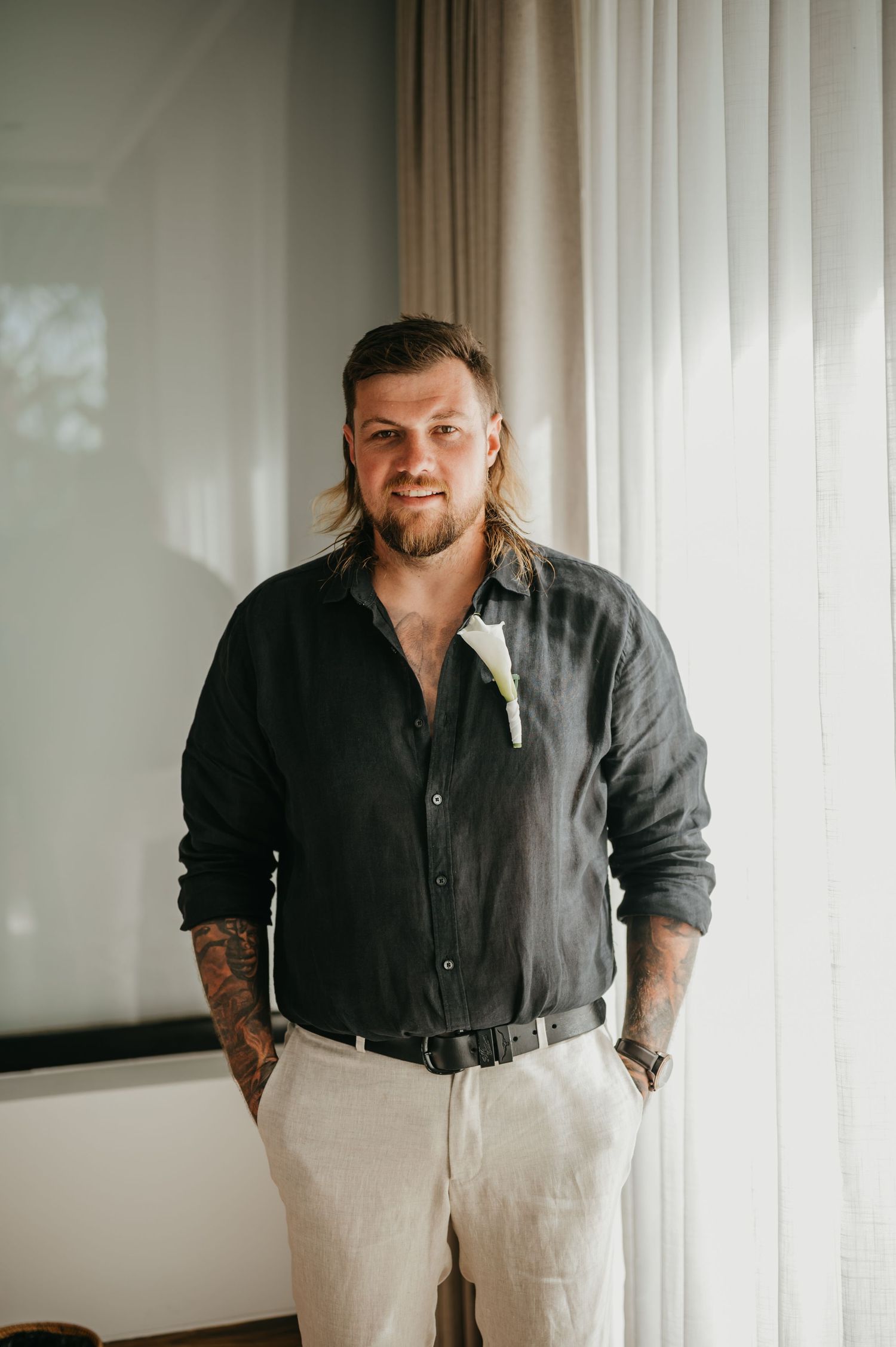 Groom in black linen shirt with floral boutonniere and tattoos posing indoors before wedding ceremony.