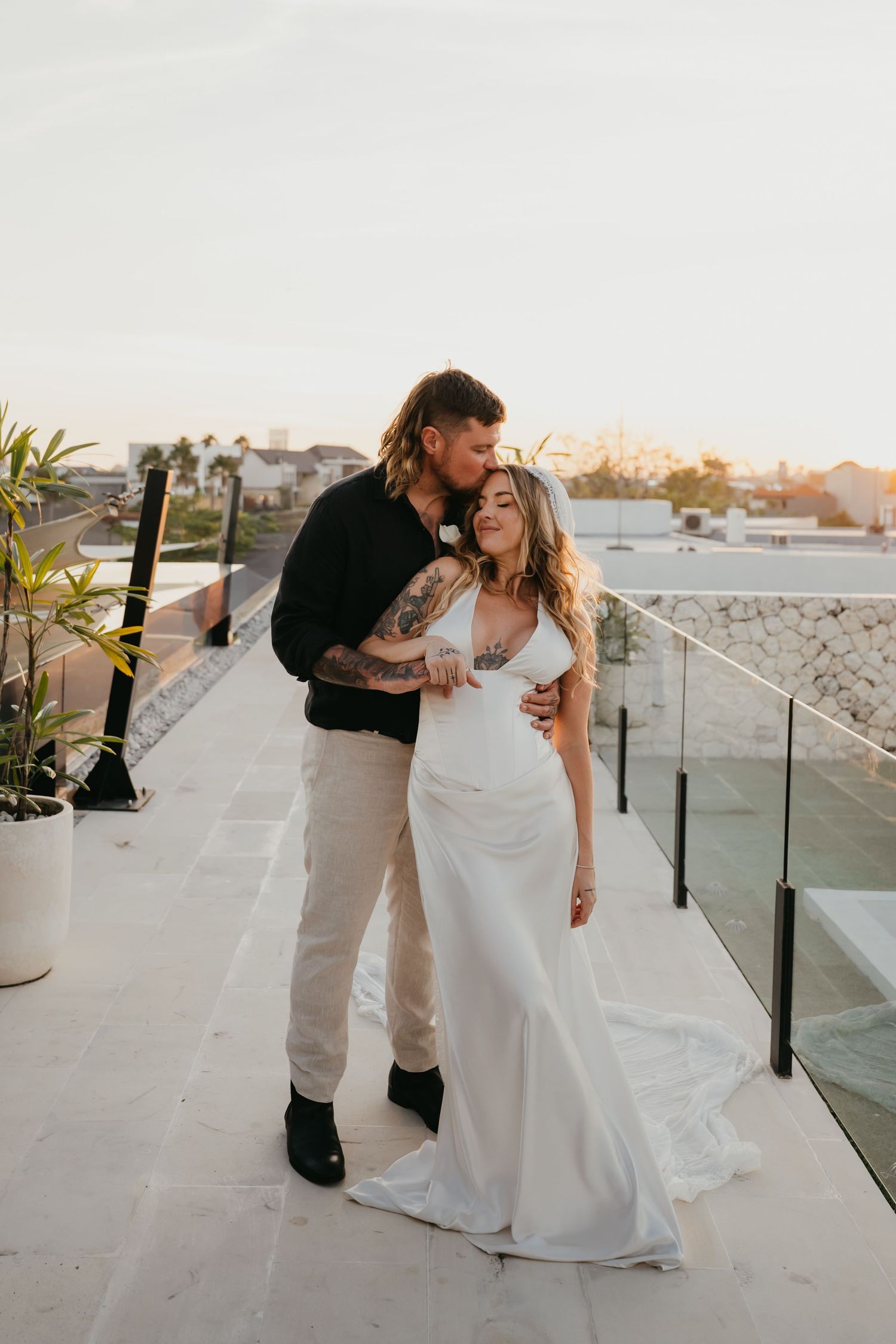 Tattooed groom in black shirt kisses bride in sleek white gown on rooftop at golden hour.