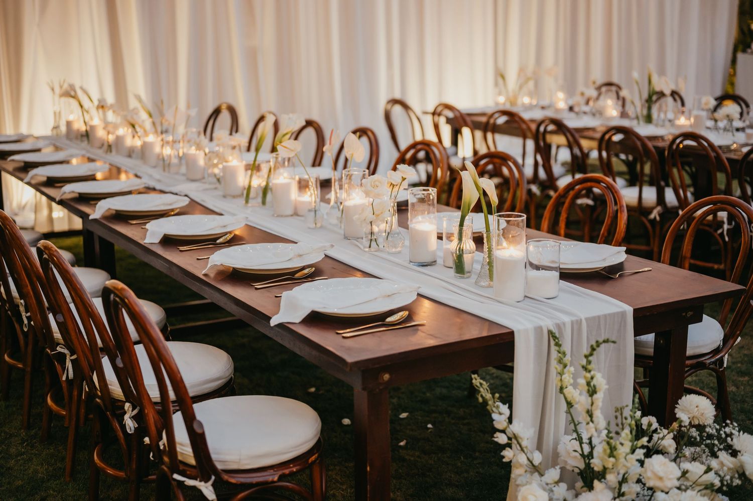 Elegant wedding reception with long wooden farm tables, white runners, candles, and bentwood chairs in a draped tent.
