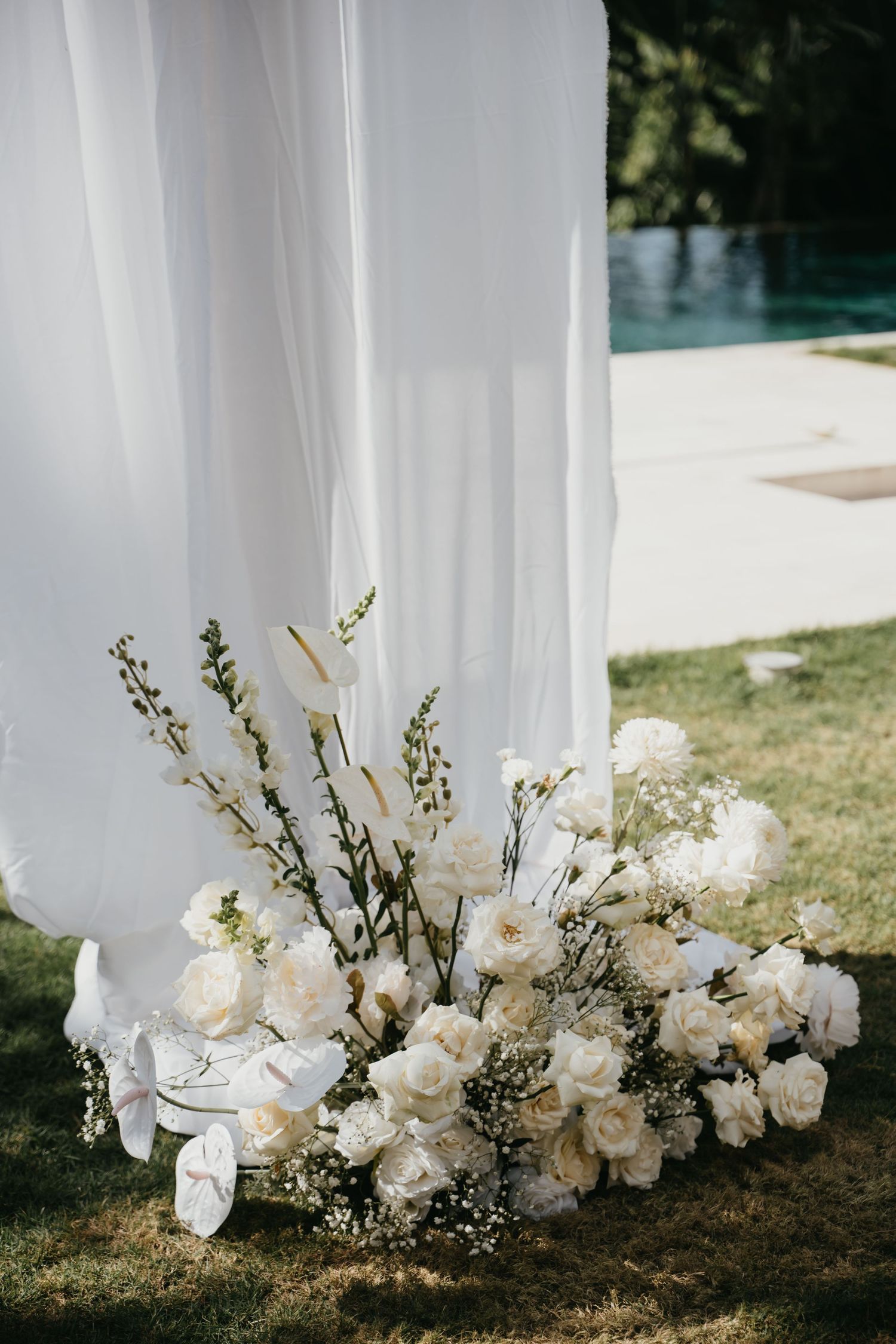 Elegant wedding arch base with white roses, baby's breath, and tall blooms against flowing white drape near a pool.