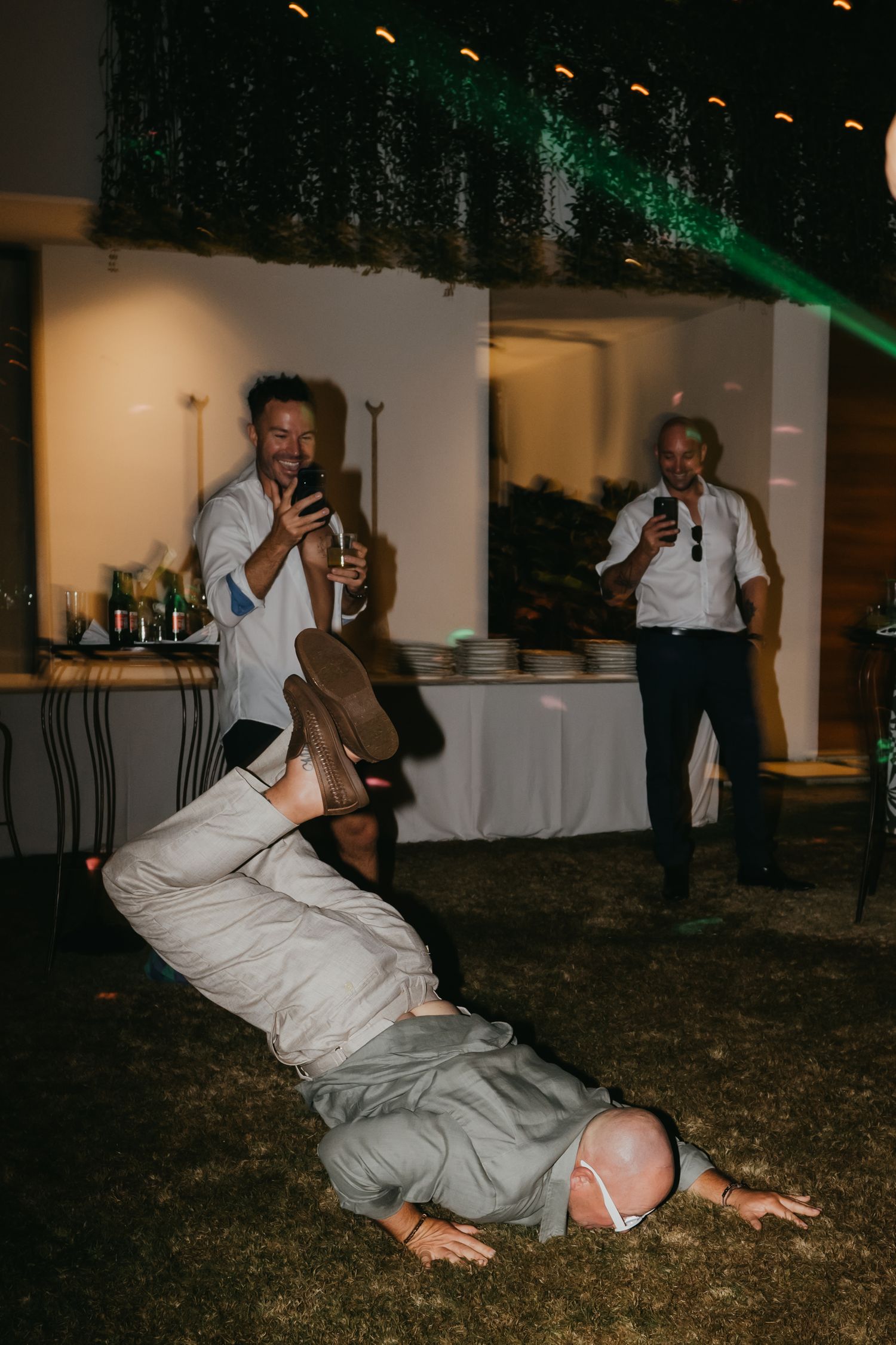 Guest breakdancing on grass while others laugh and film at outdoor wedding reception.