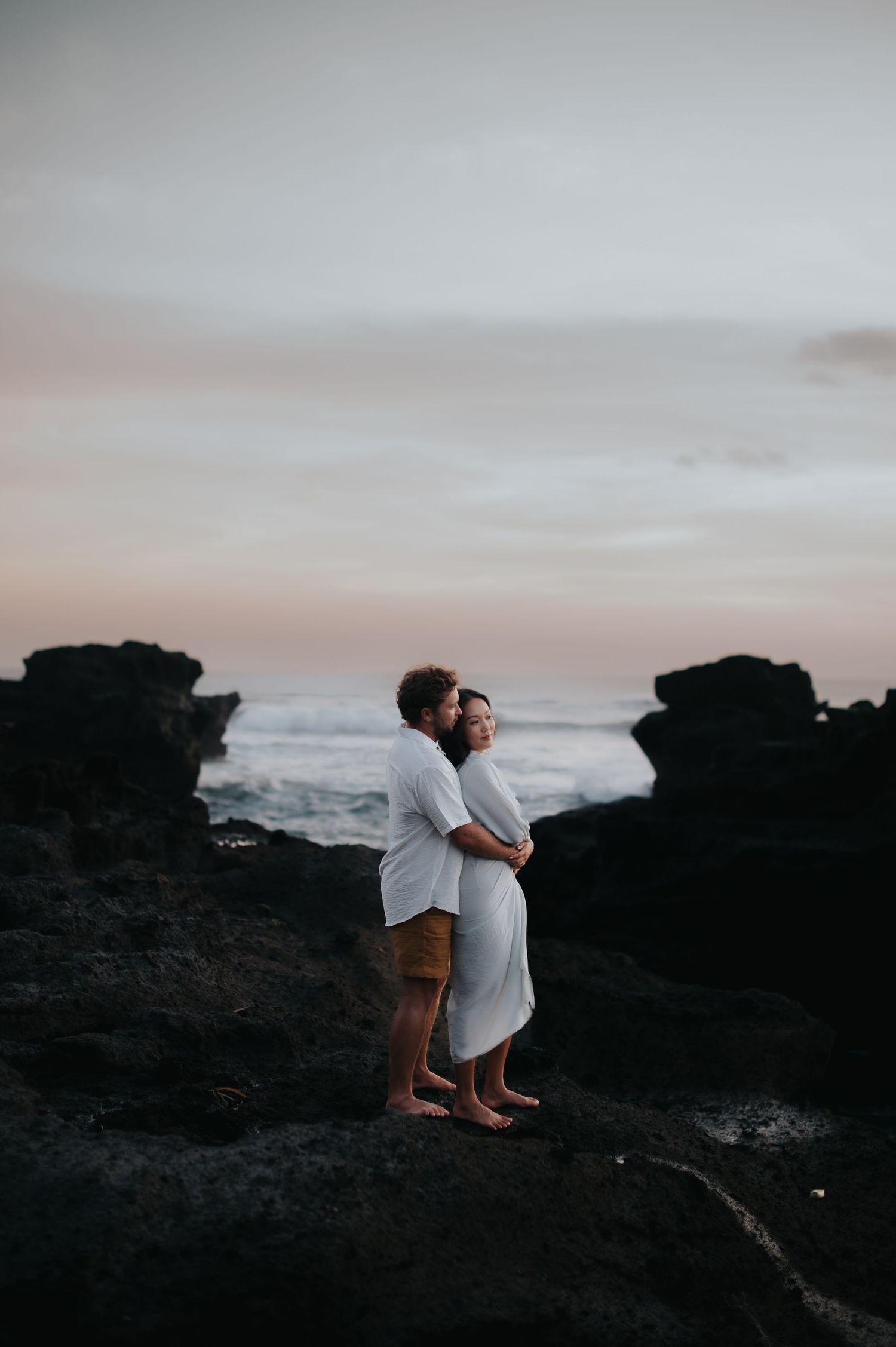 A couple embraces on dark rocky cliffs overlooking the ocean at sunset, creating a romantic silhouette against the misty sky.