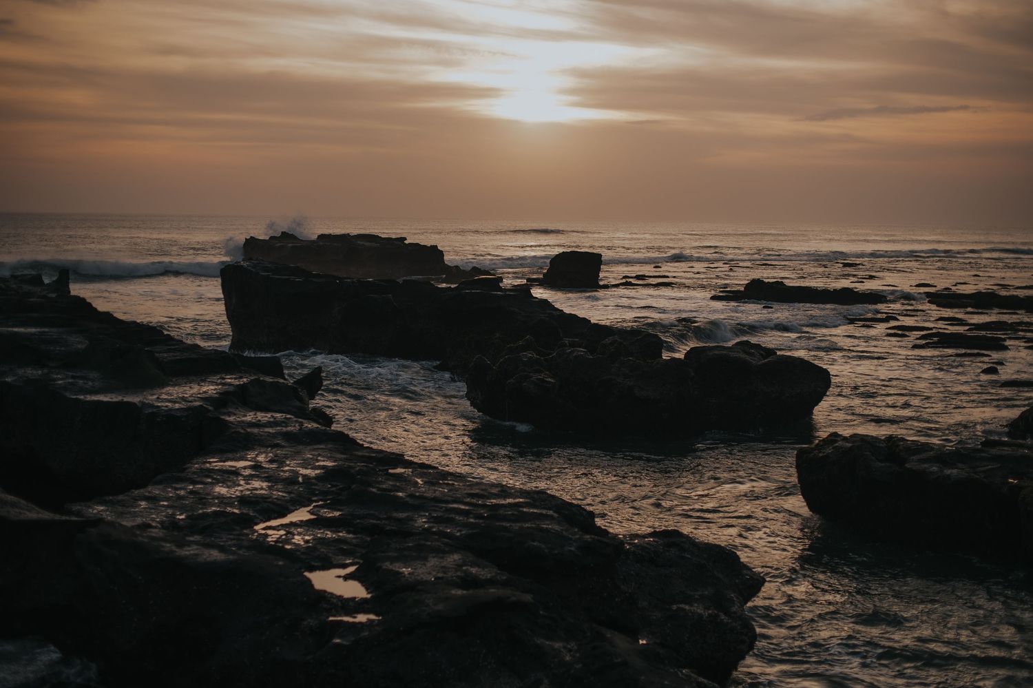 Dramatic sunset over rocky coastal landscape with dark silhouetted cliffs and crashing waves against moody sky.