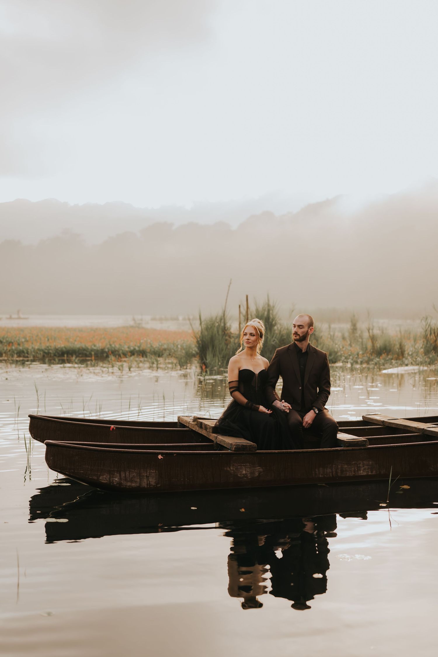 A couple sits together in a wooden canoe on a tranquil misty lake at dawn.