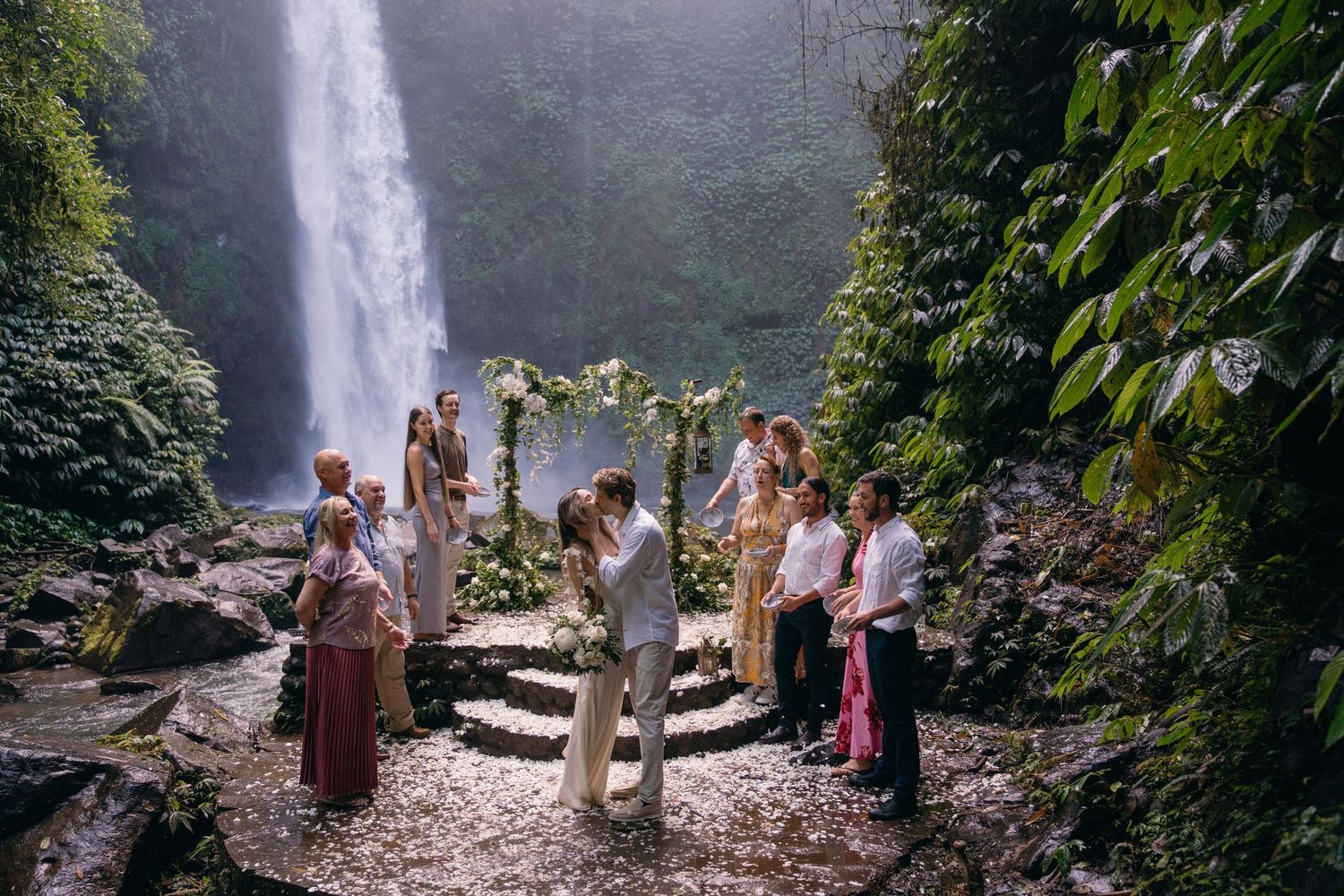 A group of tourists gather near a misty waterfall in a lush tropical forest setting.
