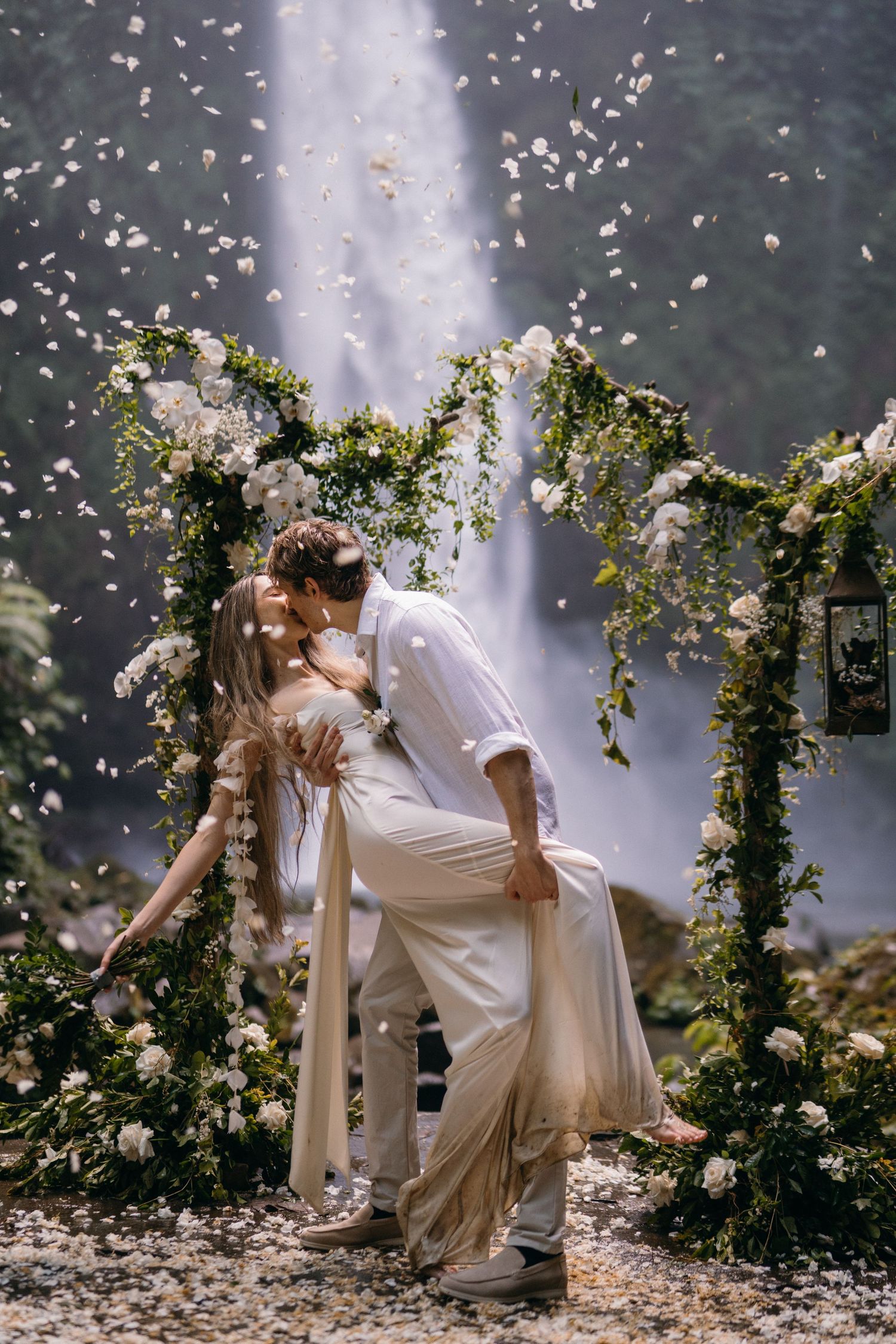 A romantic couple shares a kiss under a floral arch while white petals fall around them in a dreamy outdoor setting.