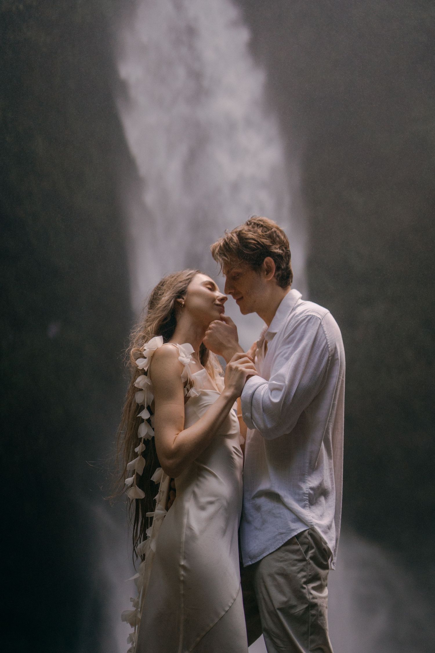 A romantic couple shares an intimate moment in front of a misty waterfall backdrop.