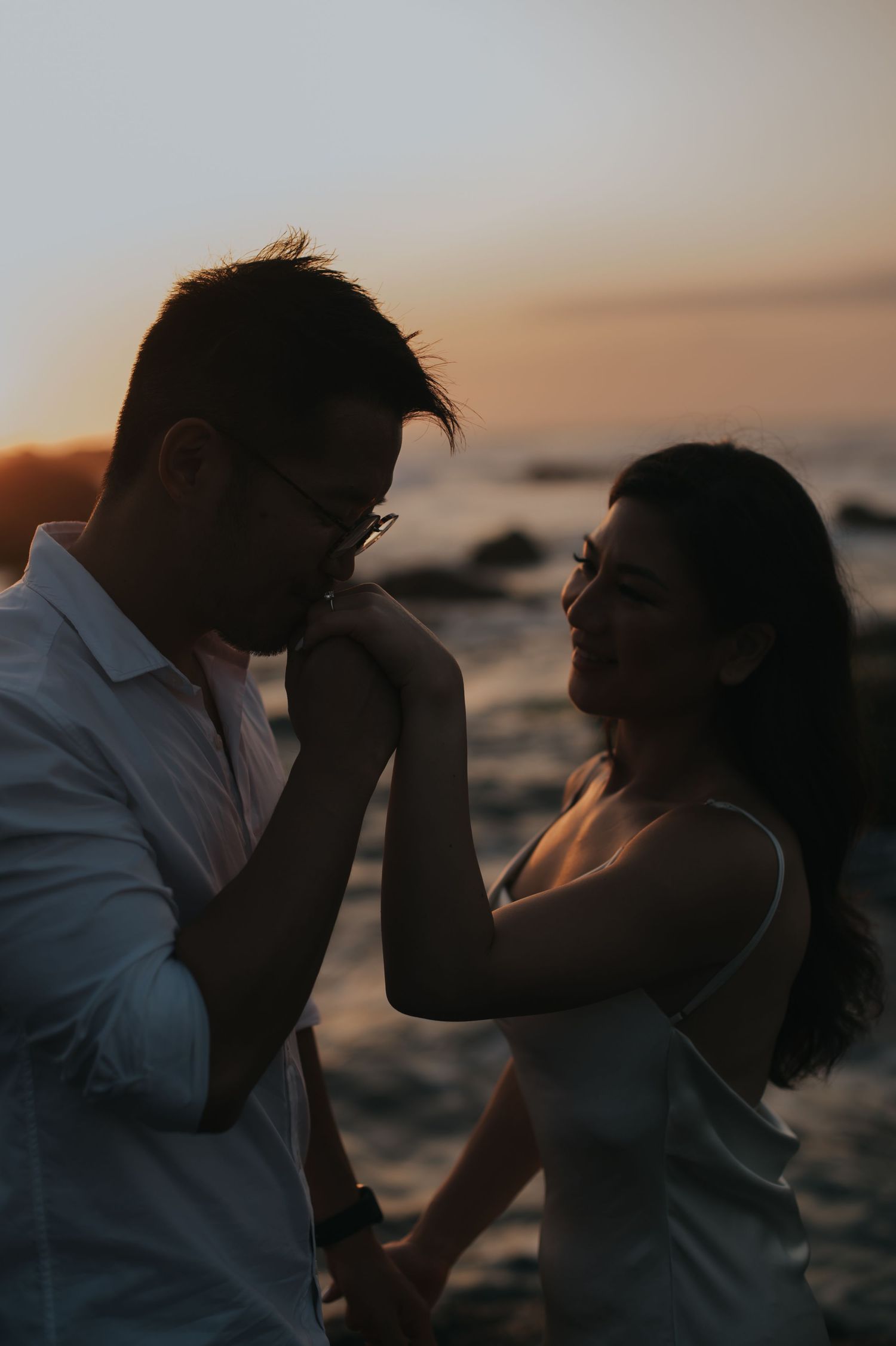 Romantic couple silhouetted against a golden sunset by the ocean, man kissing woman's hand while holding hands.