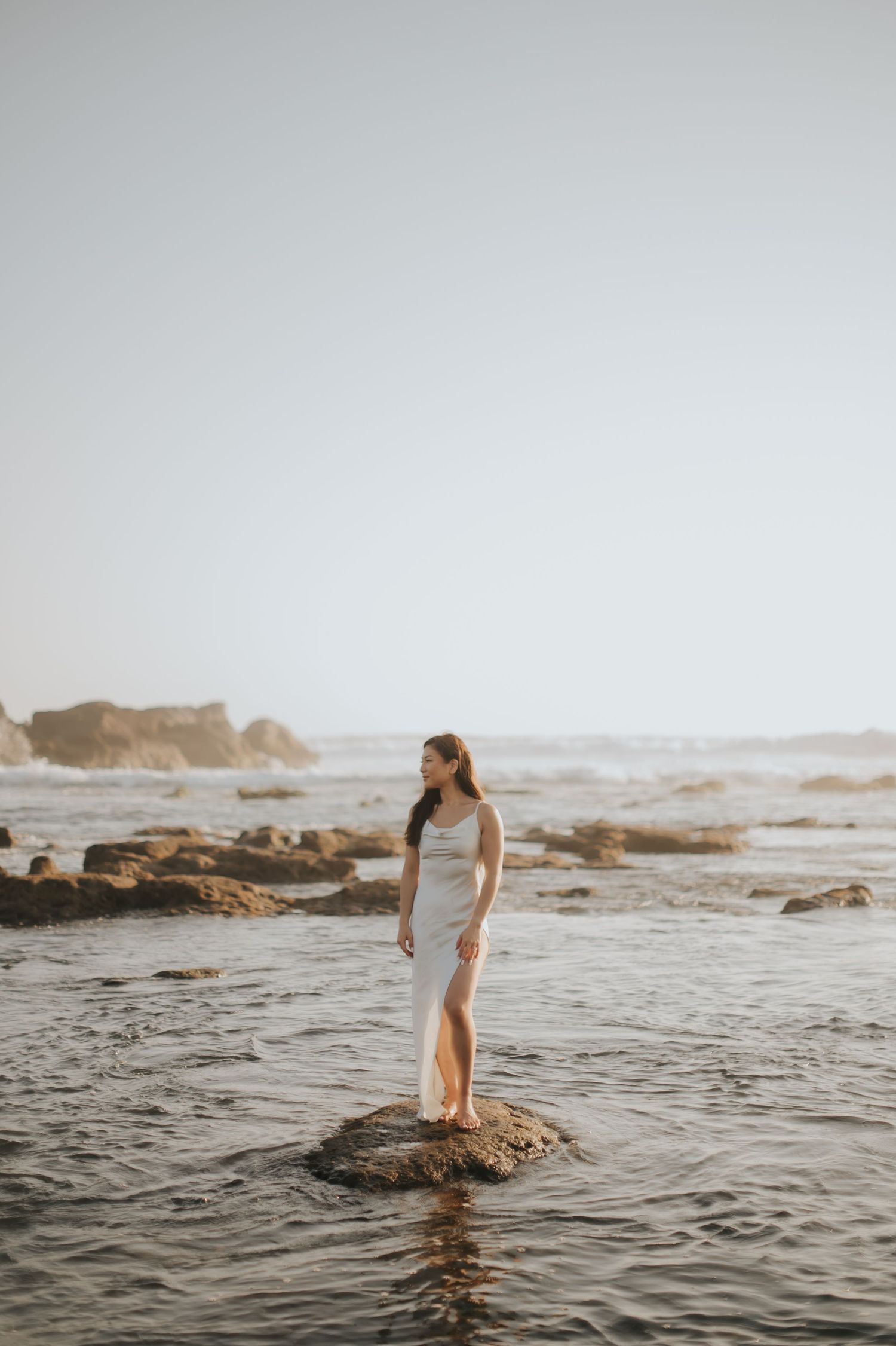 Woman in white dress standing on coastal rocks with ocean waves and misty horizon in background.