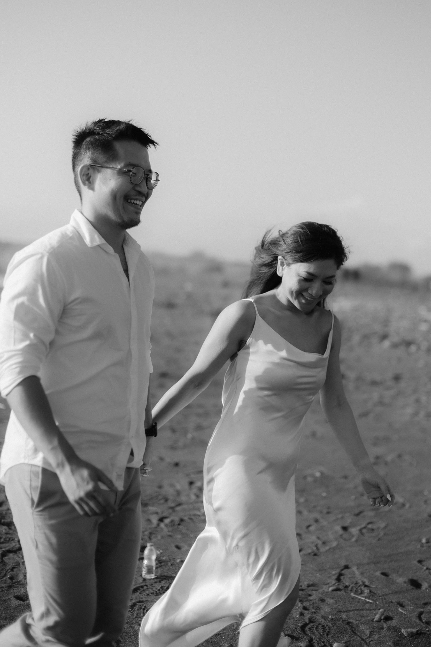 Black and white photo of a happy couple holding hands and walking together on a sandy beach in elegant attire.