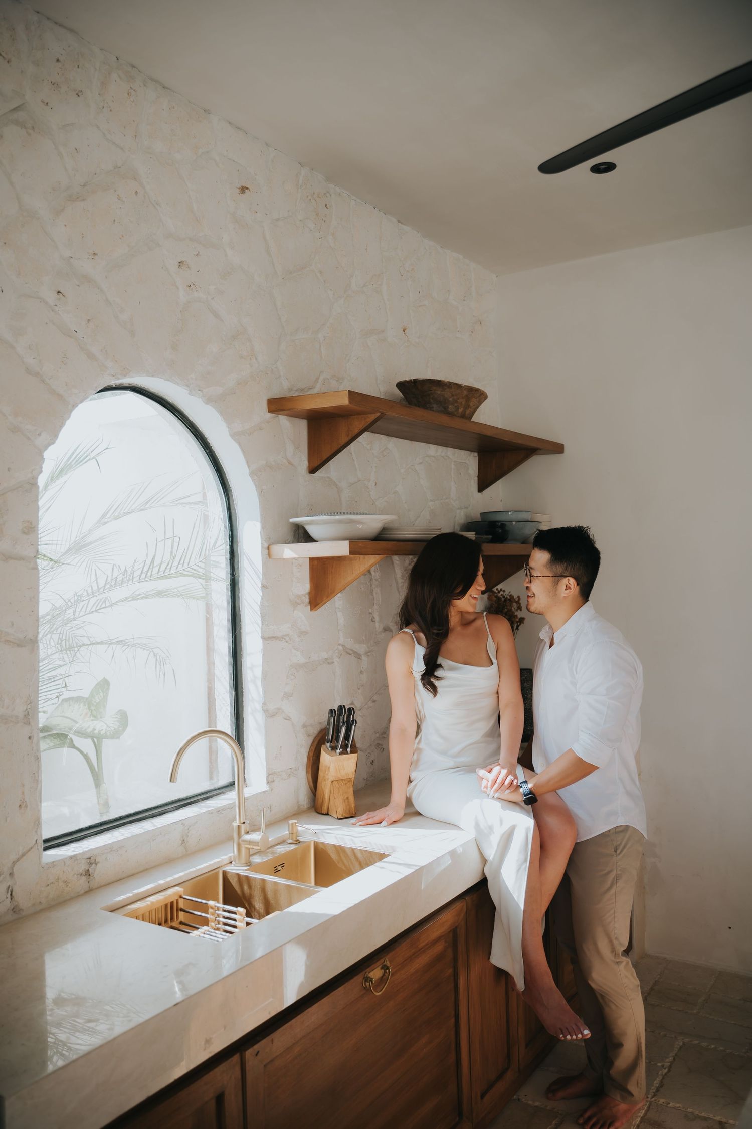 A couple shares an intimate moment in a bright boho kitchen, the woman sitting on the counter near an arched window.