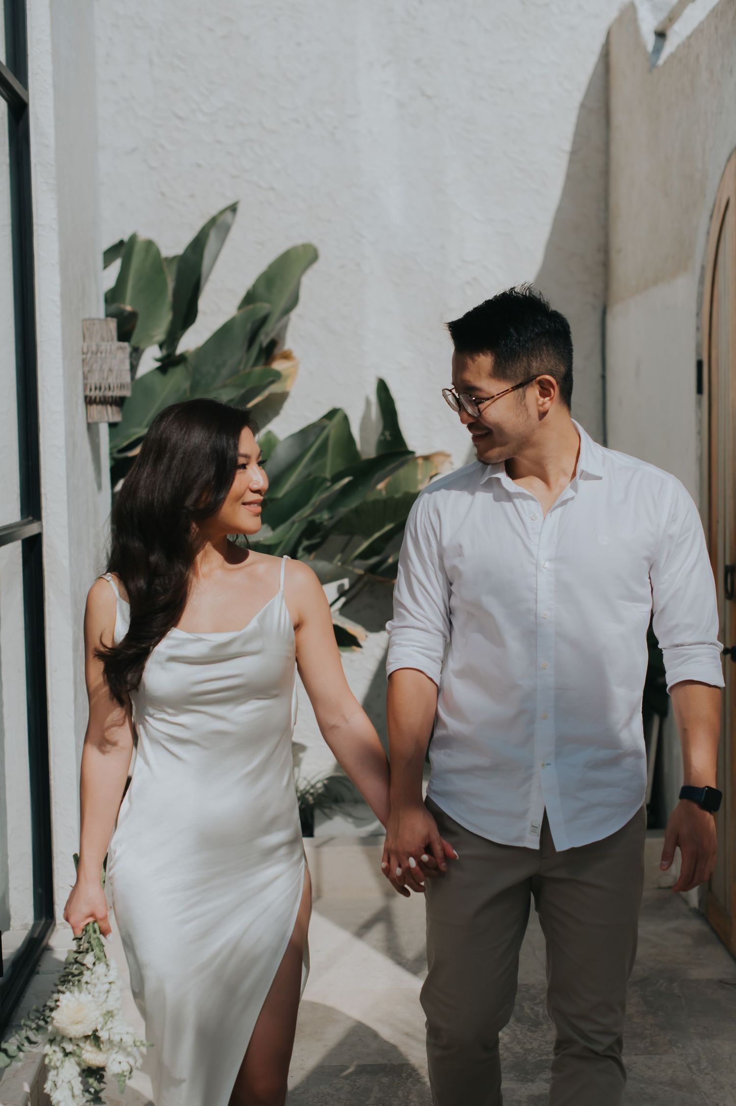Couple holding hands walking together, woman in white satin dress holding flowers, man in white shirt, outdoor setting.