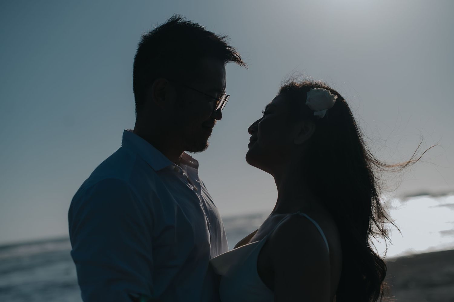 Romantic couple silhouetted against ocean sunset, woman's hair blowing in breeze, sharing intimate moment on beach.