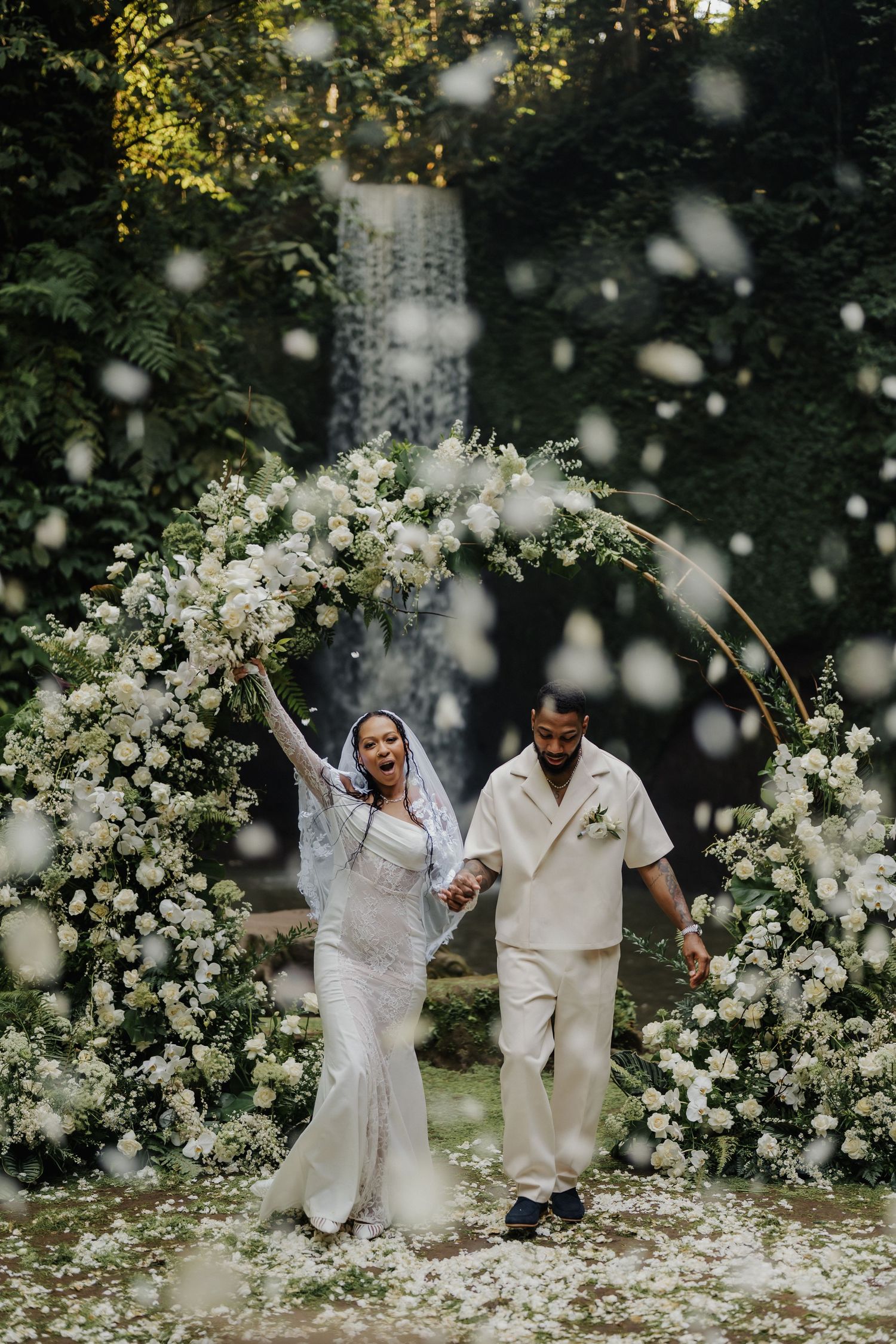 Radiant newlyweds walk through circular white floral arch as flower petals fall at lush tropical waterfall venue.