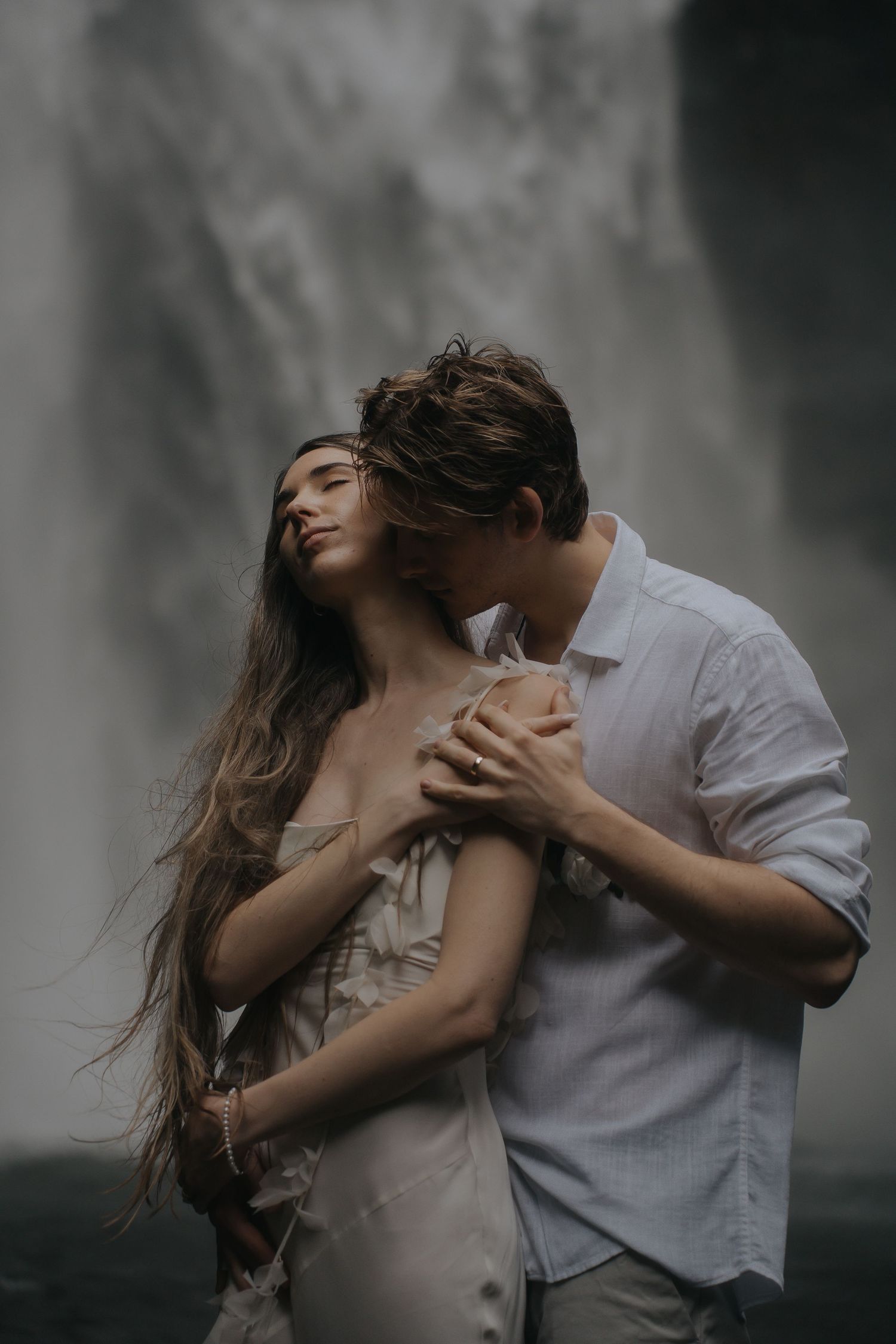 Romantic couple embracing near misty waterfall in moody atmospheric portrait.