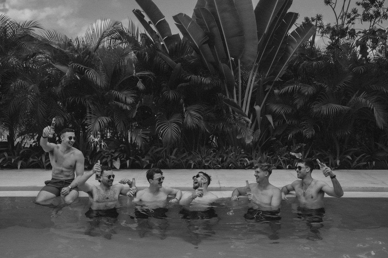 A black and white photograph shows people relaxing in a swimming pool surrounded by tropical palm trees and foliage.
