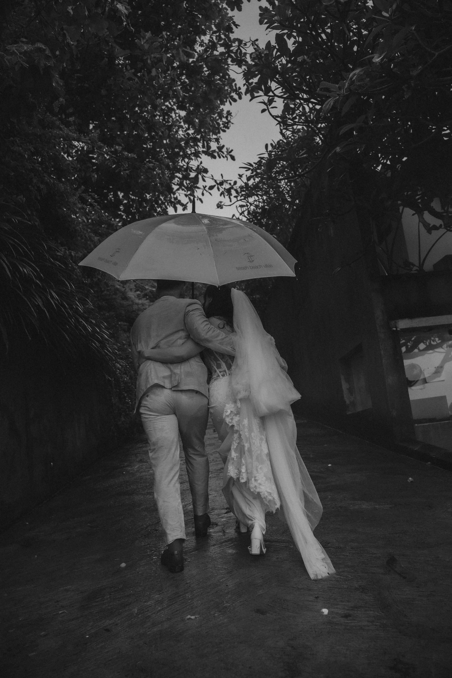 Two people walking together under umbrellas in the rain along a dark path through trees.