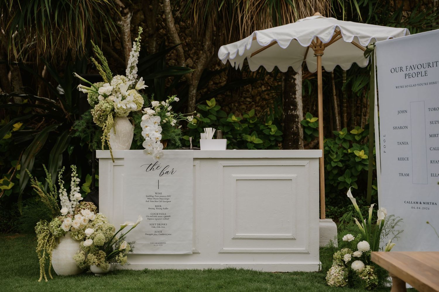 Elegant white outdoor wedding bar setup with floral arrangements and decorative umbrella in garden.