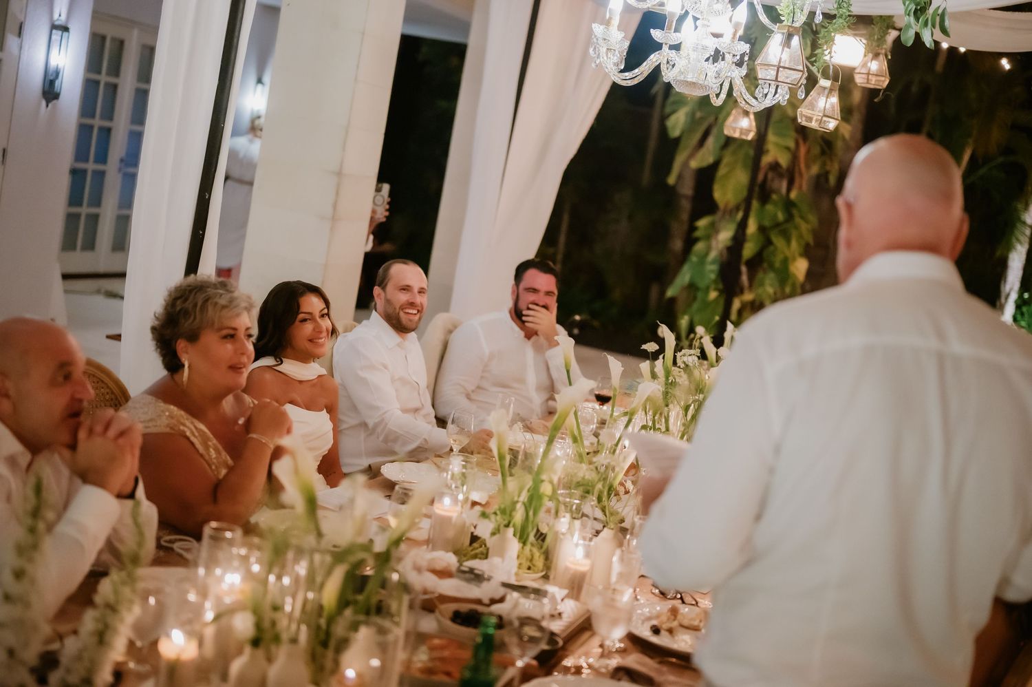 Wedding guests and couple laugh during heartfelt speech at candlelit reception with floral centerpieces.