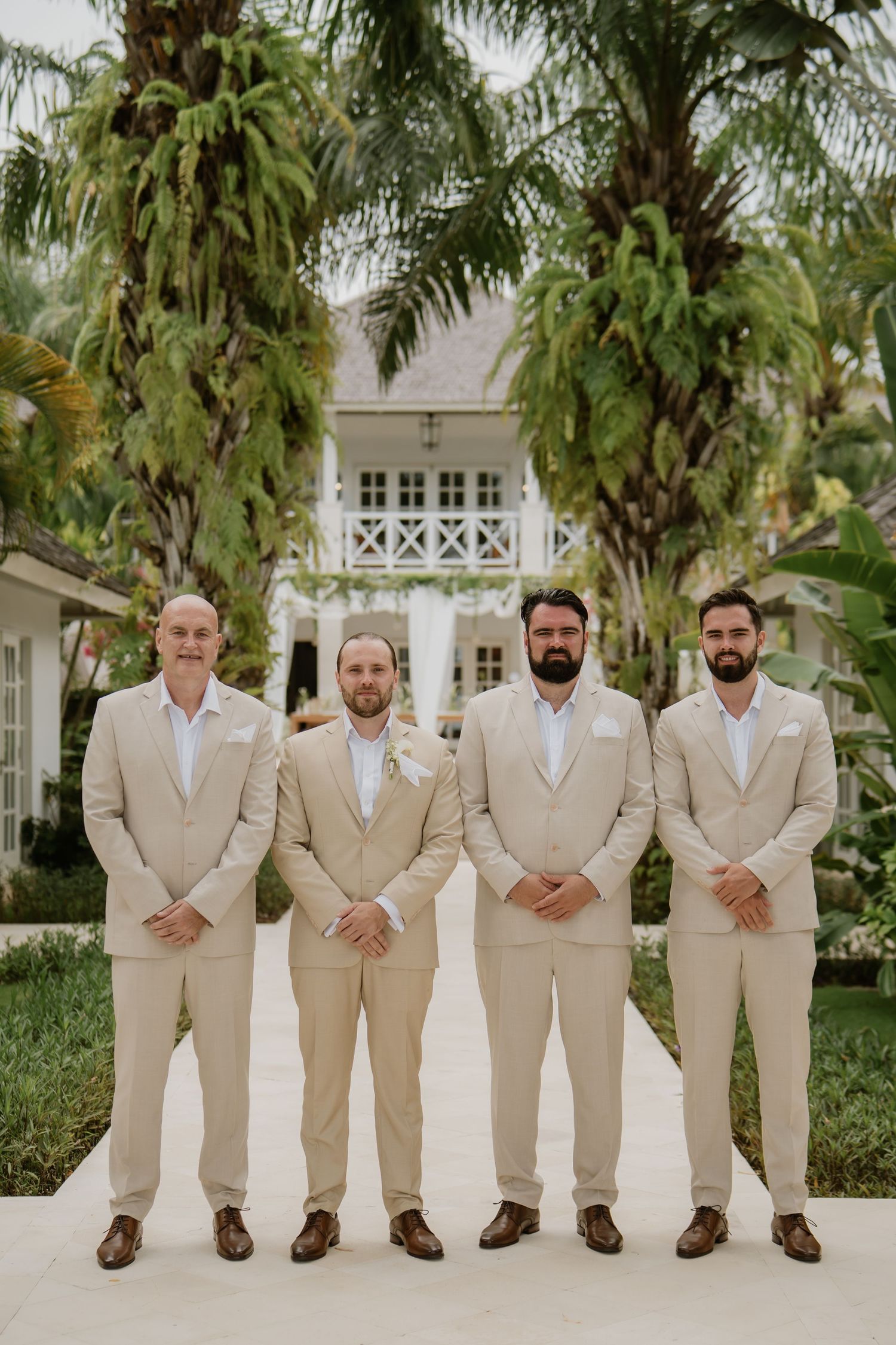 Four groomsmen in matching beige suits pose outdoors in front of a tropical estate.