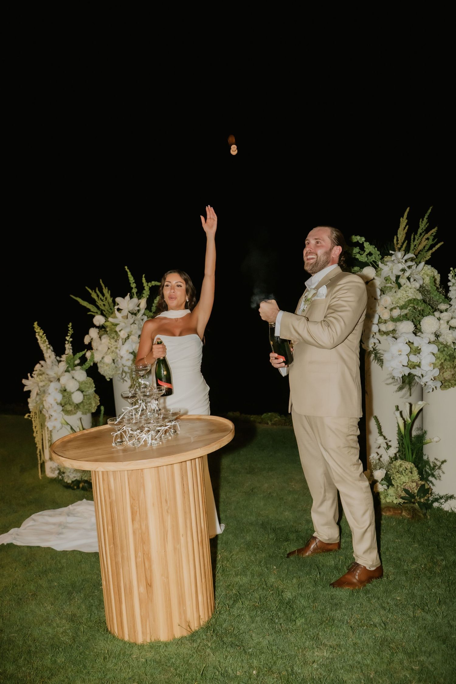 Excited bride and groom popping champagne bottle at night in front of white floral wedding arrangements.