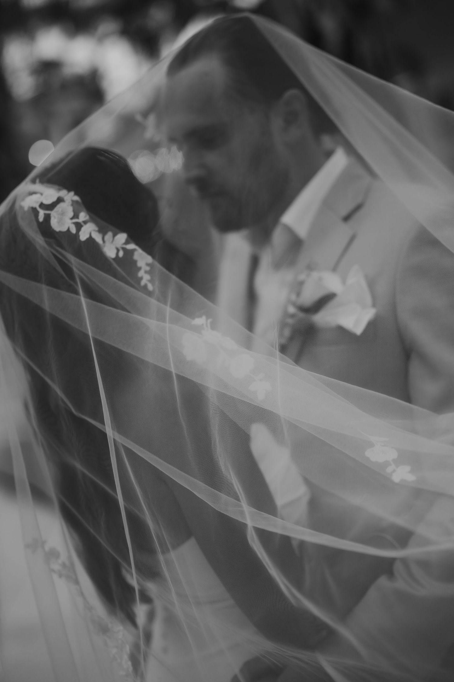 Black and white romantic photo of bride and groom sharing intimate moment beneath flowing lace veil.