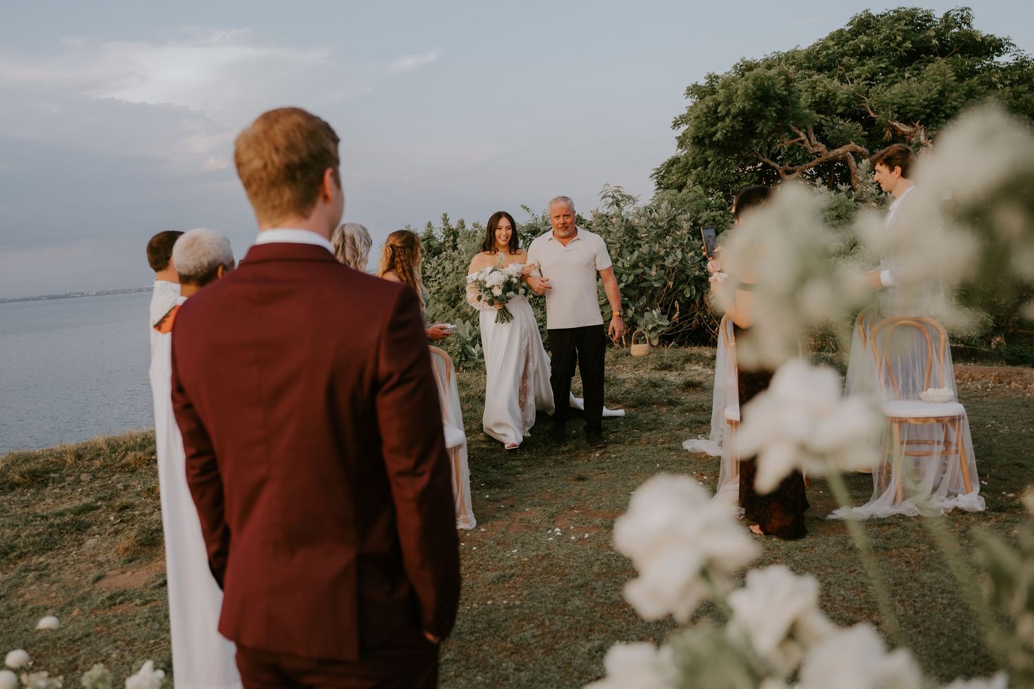 Bride walks down the aisle toward groom at a scenic outdoor cliffside wedding ceremony at sunset.