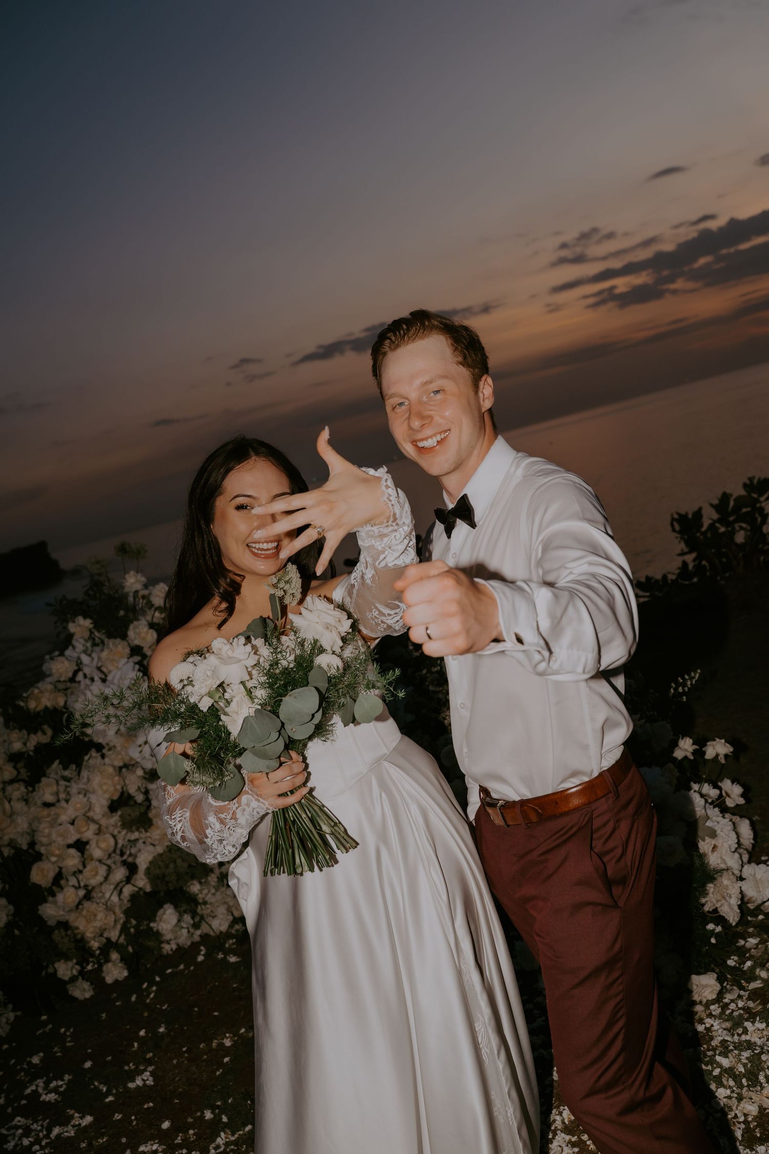 Newlyweds joyfully show off wedding rings at sunset outdoor celebration with white bouquet.