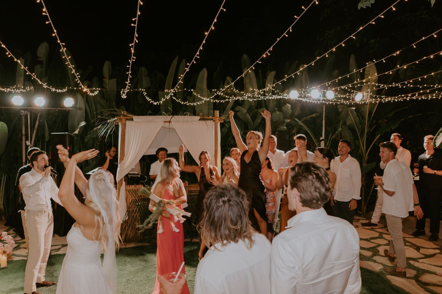 Wedding guests cheering and dancing with the bride and groom at a festive outdoor tropical night reception.