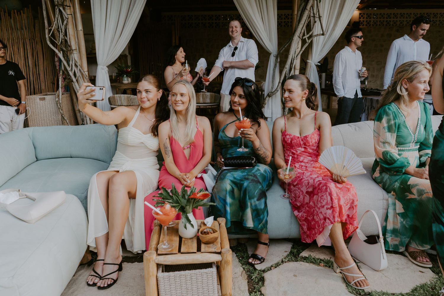 Wedding guests in colorful dresses sitting on a sofa taking a selfie at an outdoor reception.