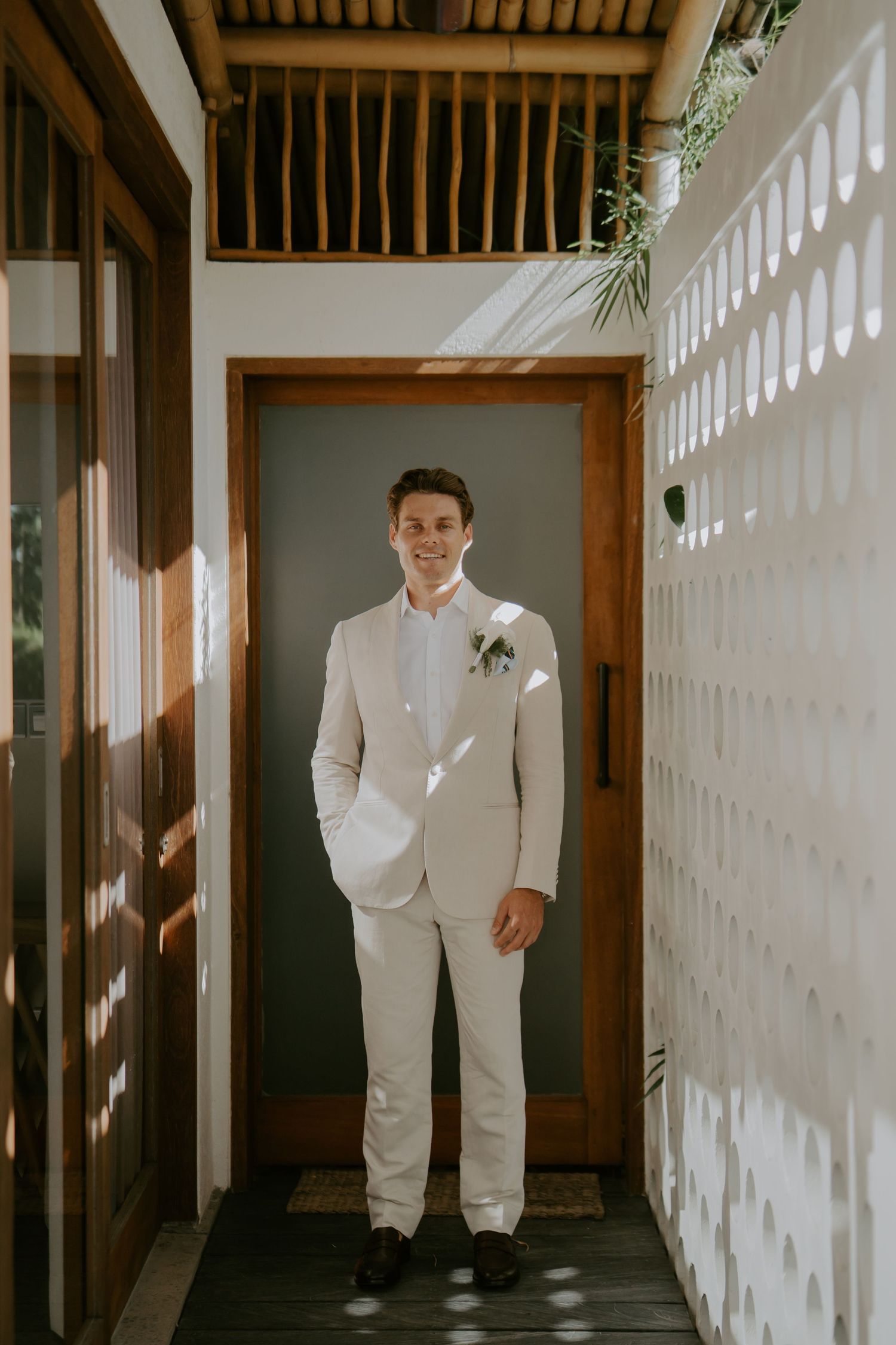 Groom in a beige suit with boutonniere stands smiling in a tropical venue hallway.