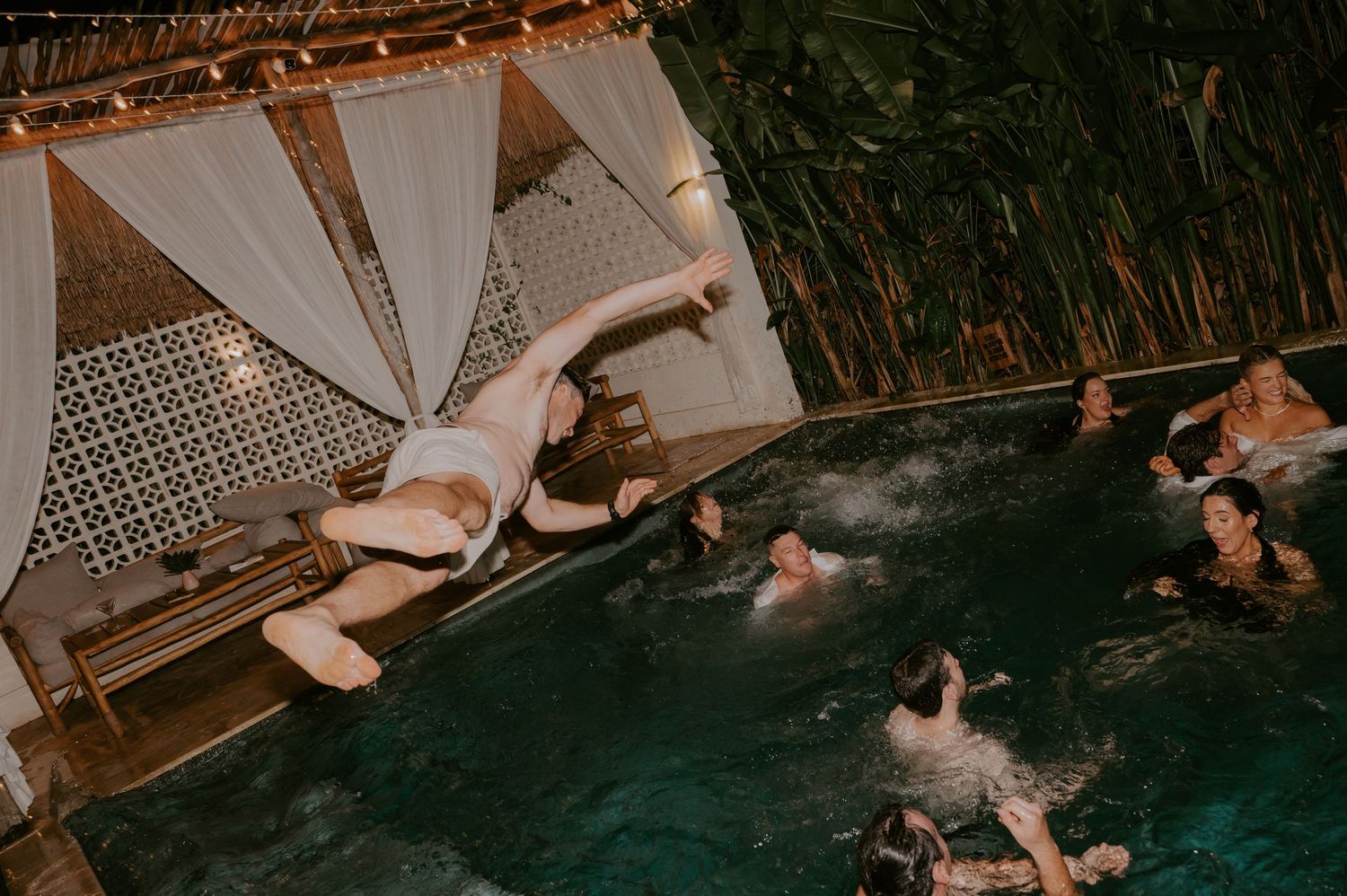 Wedding guest dramatically diving into a pool while guests swim and celebrate at an outdoor tropical wedding party.