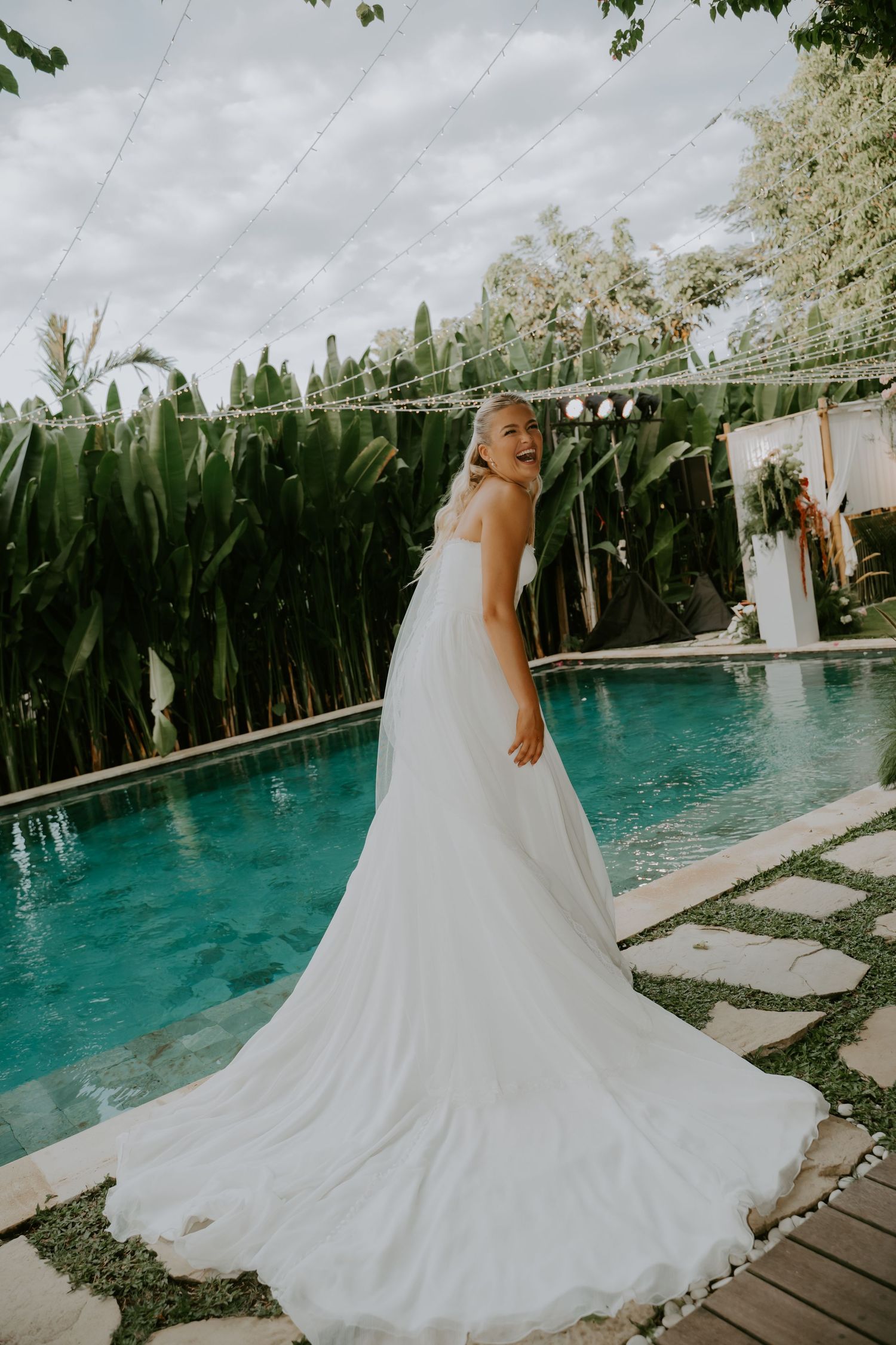 Bride in flowing white gown with long train poses beside tropical pool surrounded by lush greenery.