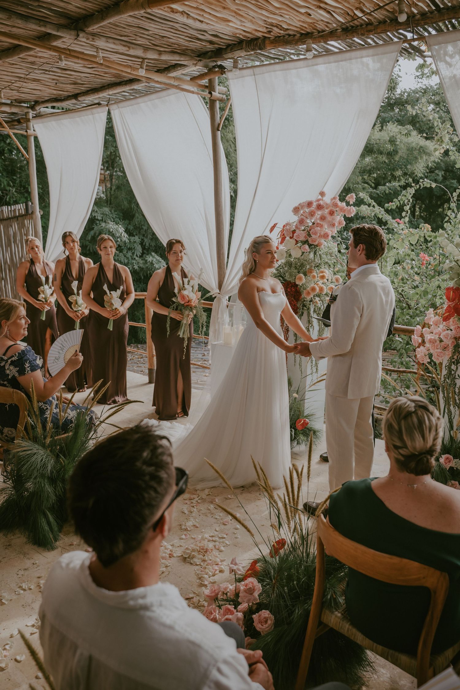 Bride and groom exchange vows under a bamboo canopy adorned with white drapes and pink floral arrangements.