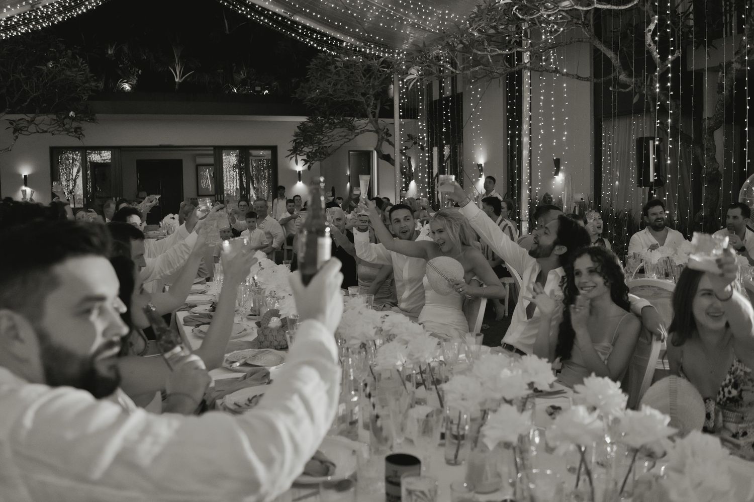 Black and white photo of wedding guests raising glasses in a toast at reception dinner.