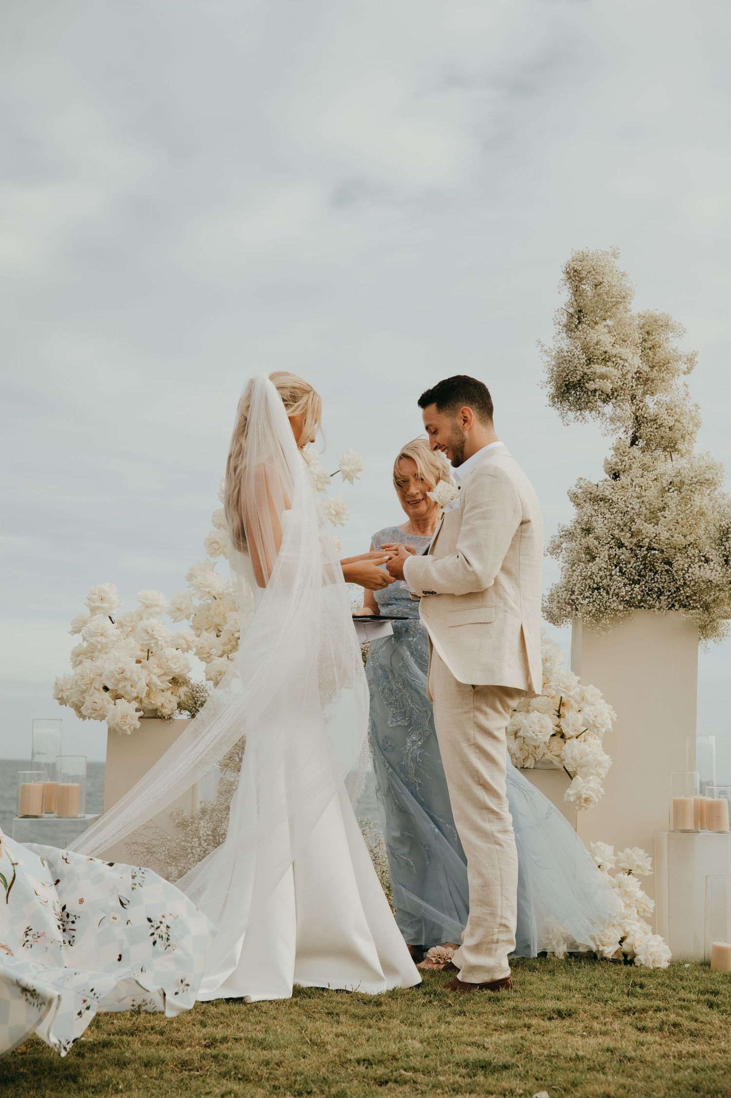 Bride and groom exchange rings at oceanside ceremony with white floral pedestals and baby's breath backdrop.