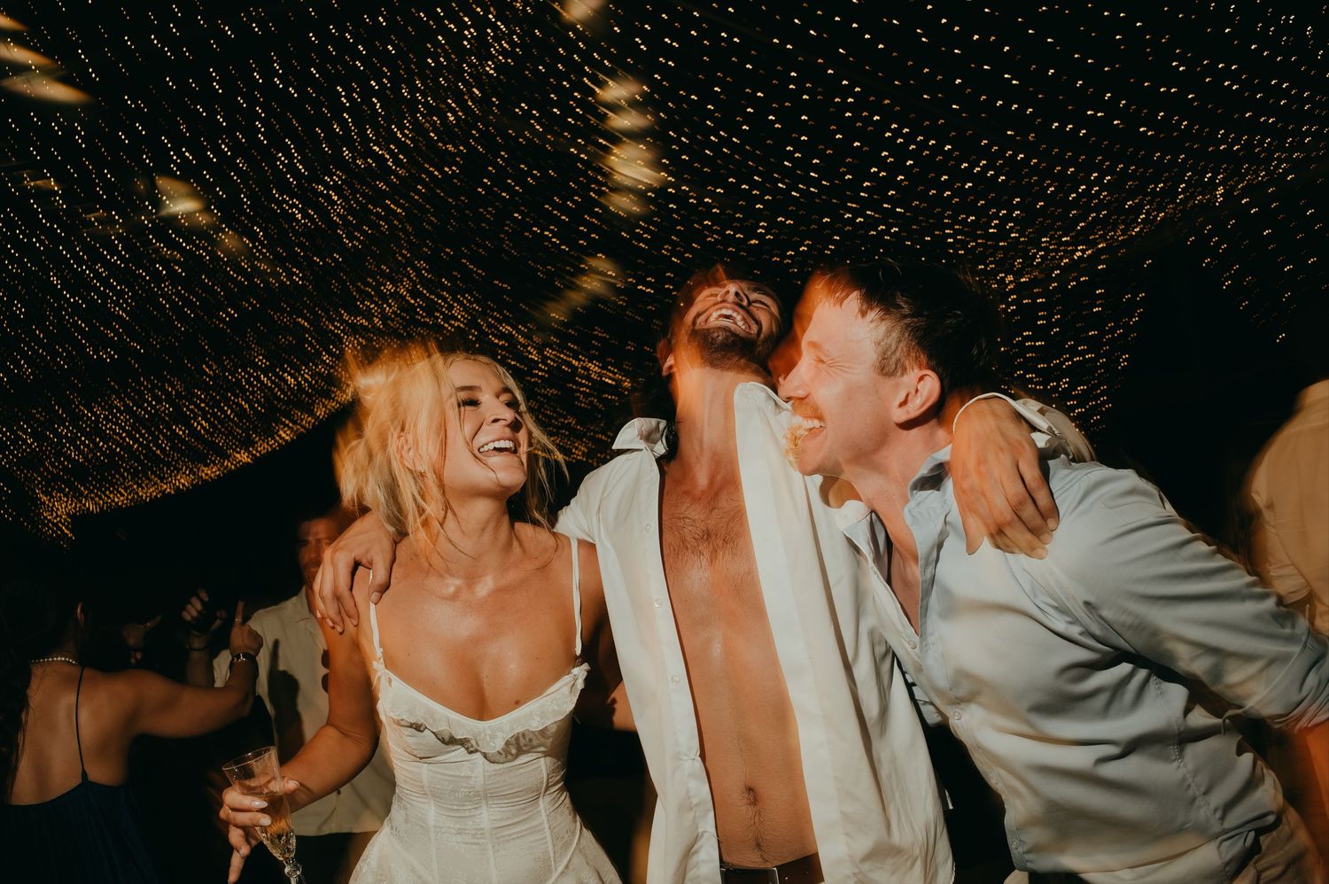 Bride and two guests laughing joyfully together on dance floor at outdoor wedding reception at night.