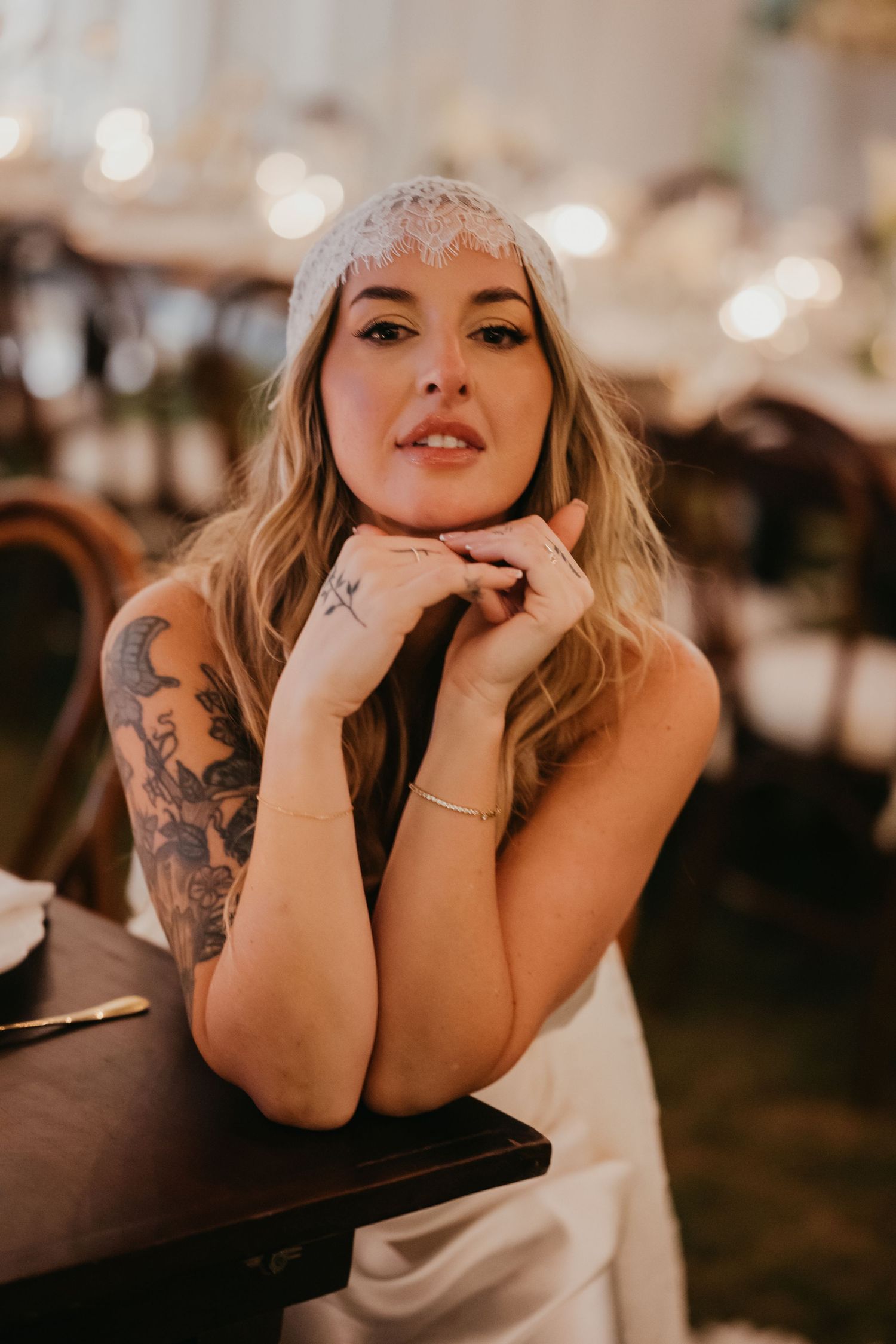 Tattooed bride wearing lace headpiece poses at wedding reception table with bokeh light background.
