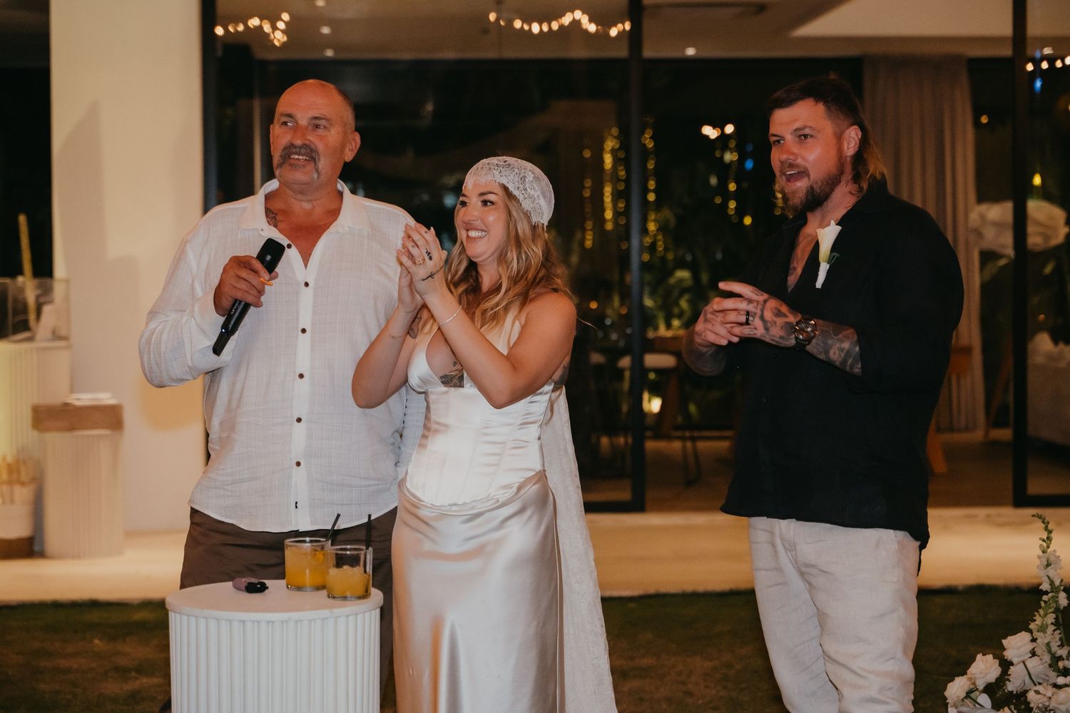 Bride and groom smile during wedding speeches with a guest holding a microphone at their reception.