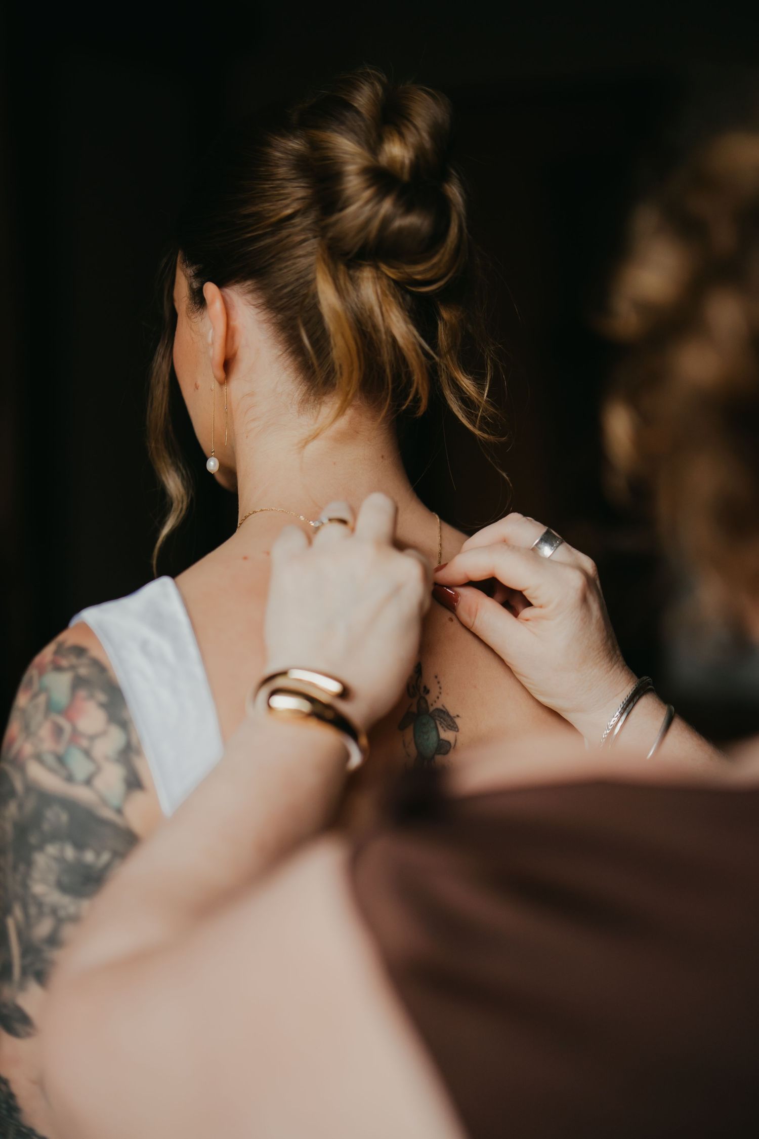 Bridesmaid fastening a delicate gold necklace on bride with updo hairstyle and floral tattoo sleeve.
