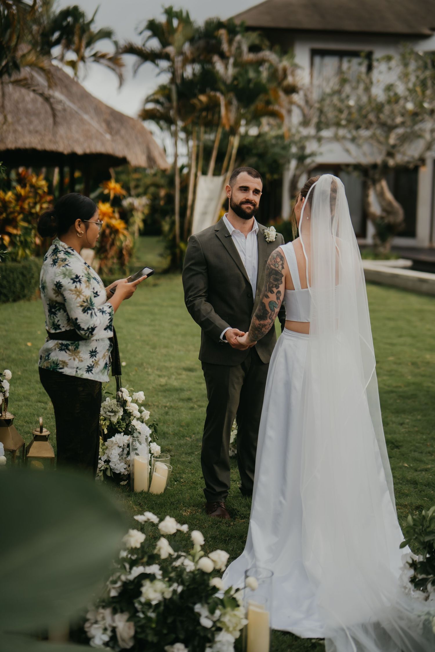 Groom in dark suit holds brides hands during outdoor wedding ceremony beside floral arrangements and palm trees.