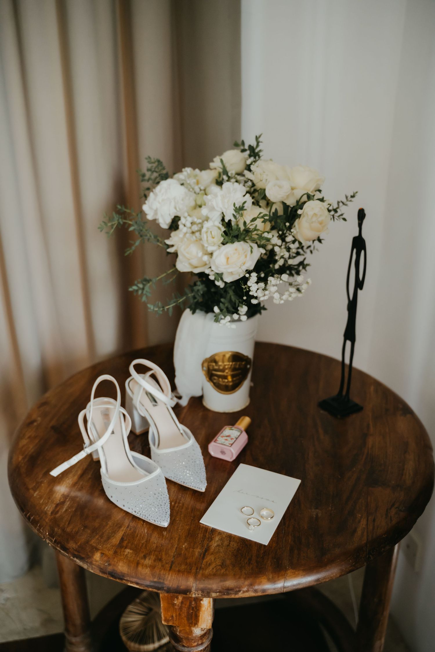 Bridal flat lay with white heels, bouquet of white roses, perfume, rings, and a card on a wooden table.