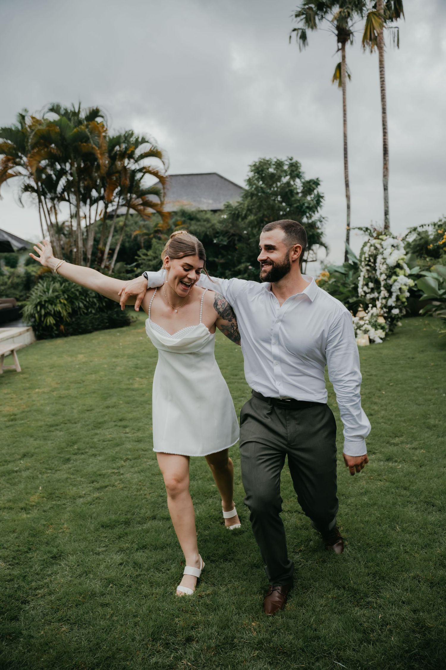 Laughing newlyweds walk arm in arm across green lawn with palm trees in background.