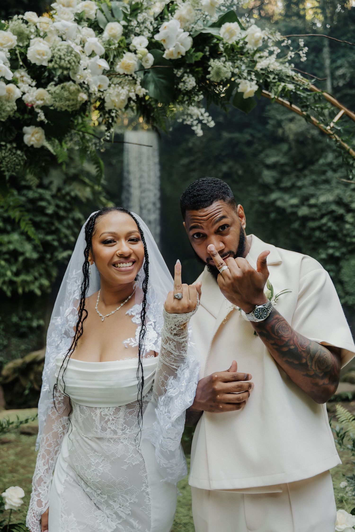 Newlywed couple showing off wedding rings under white floral arch, bride in lace veil and groom in cream suit.
