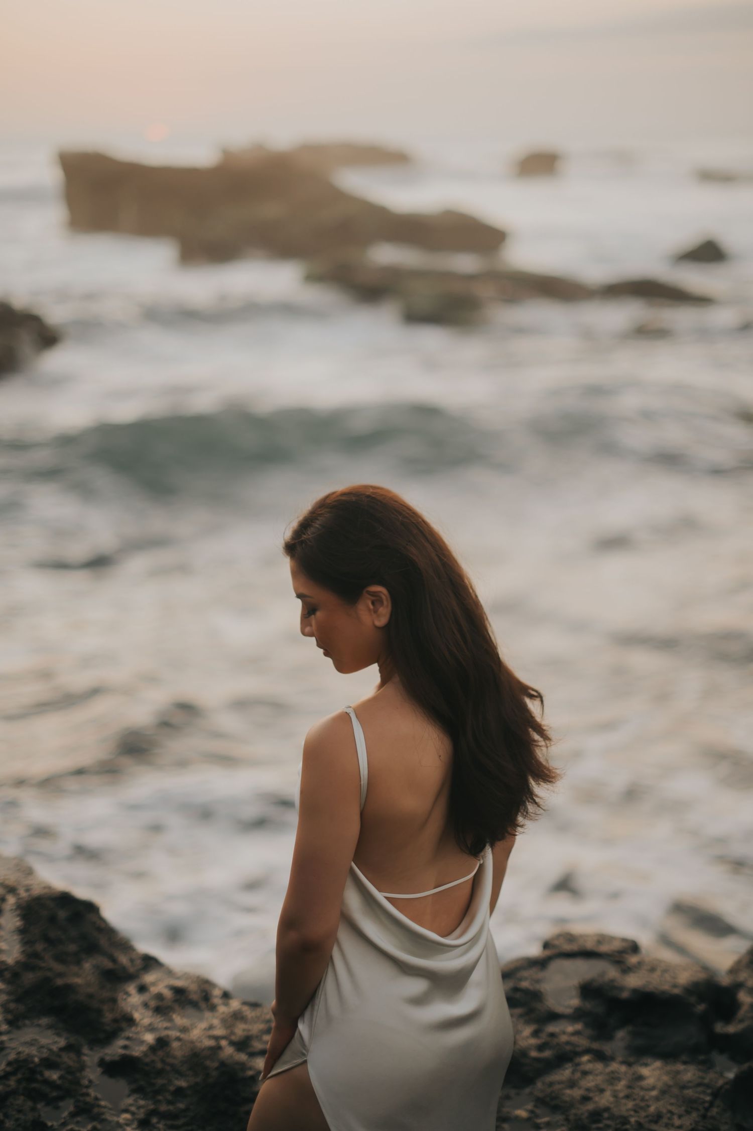 Woman in white backless dress standing on rocky shore at sunset with ocean waves in background.