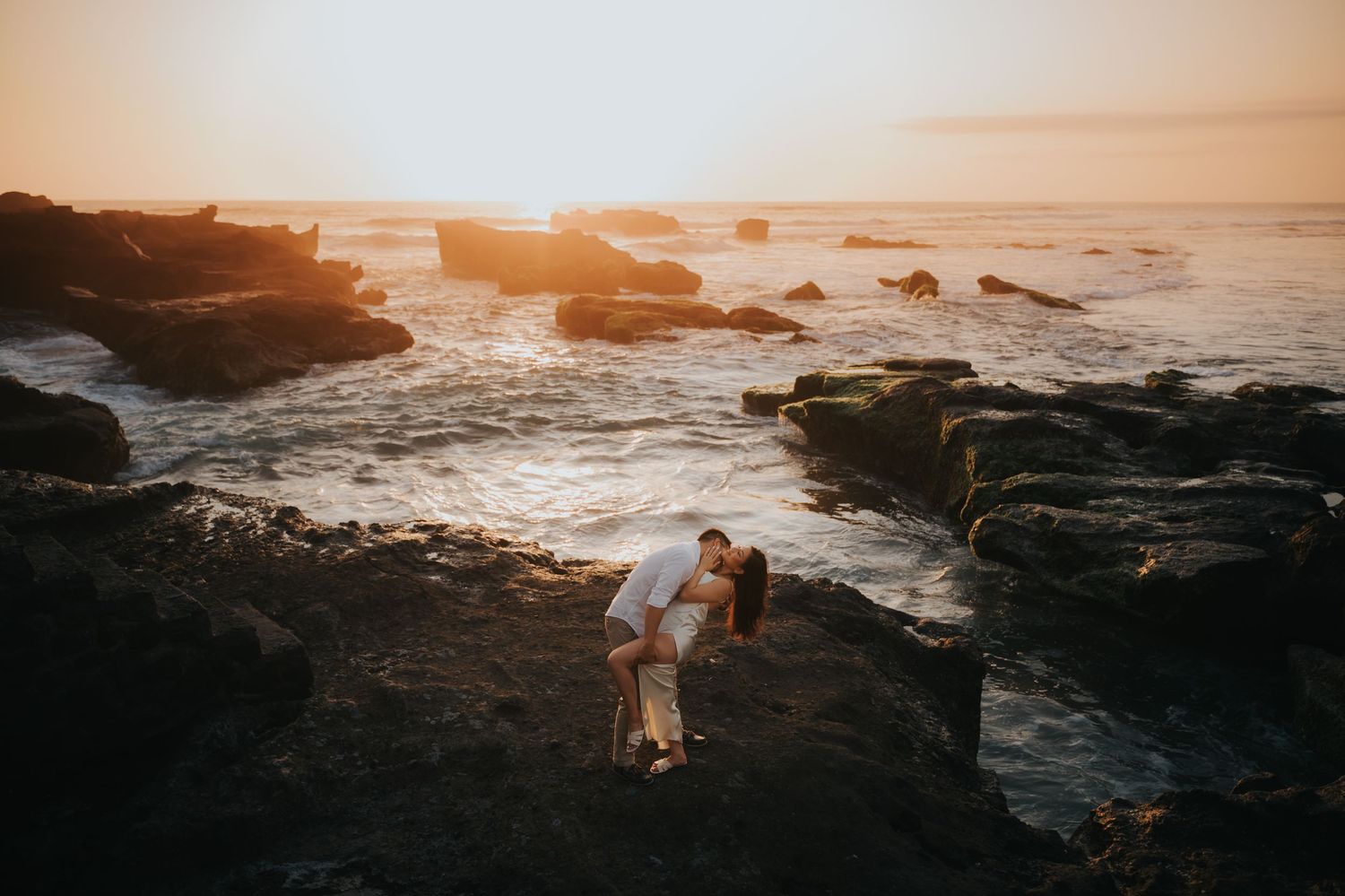 Couple sharing a romantic kiss on rocky coastal cliffs during a golden sunset with ocean waves in the background.