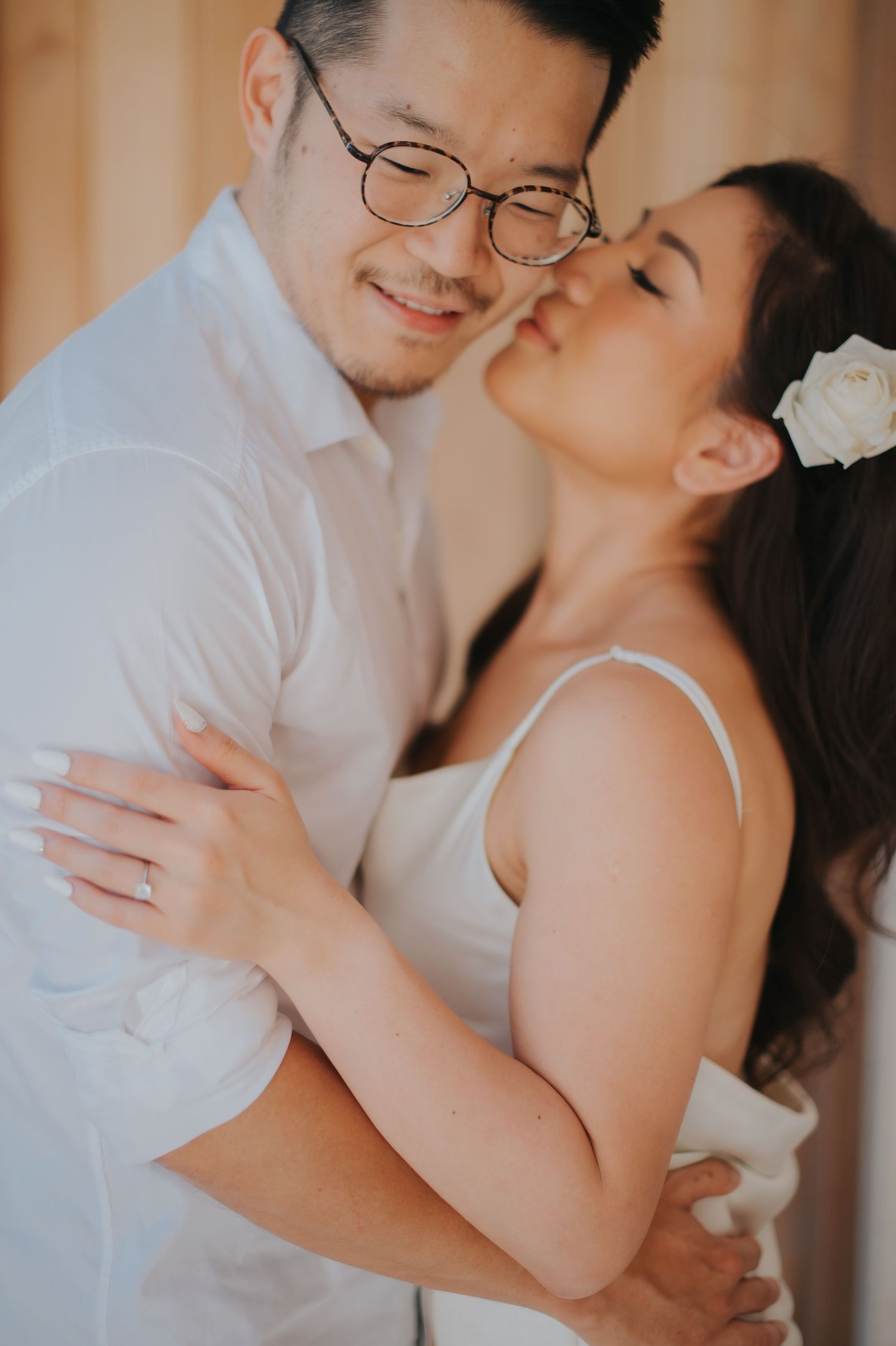 A couple in white attire shares a tender embrace, the woman kissing the man's cheek as he smiles softly.