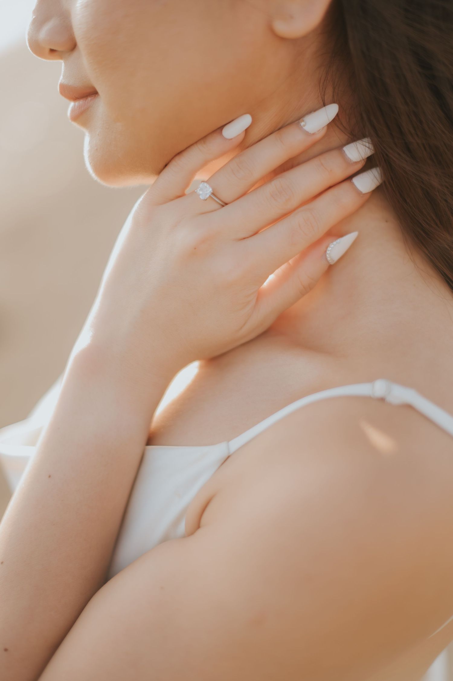 Woman touching her neck with white almond nails and a delicate gold ring, wearing a white dress.