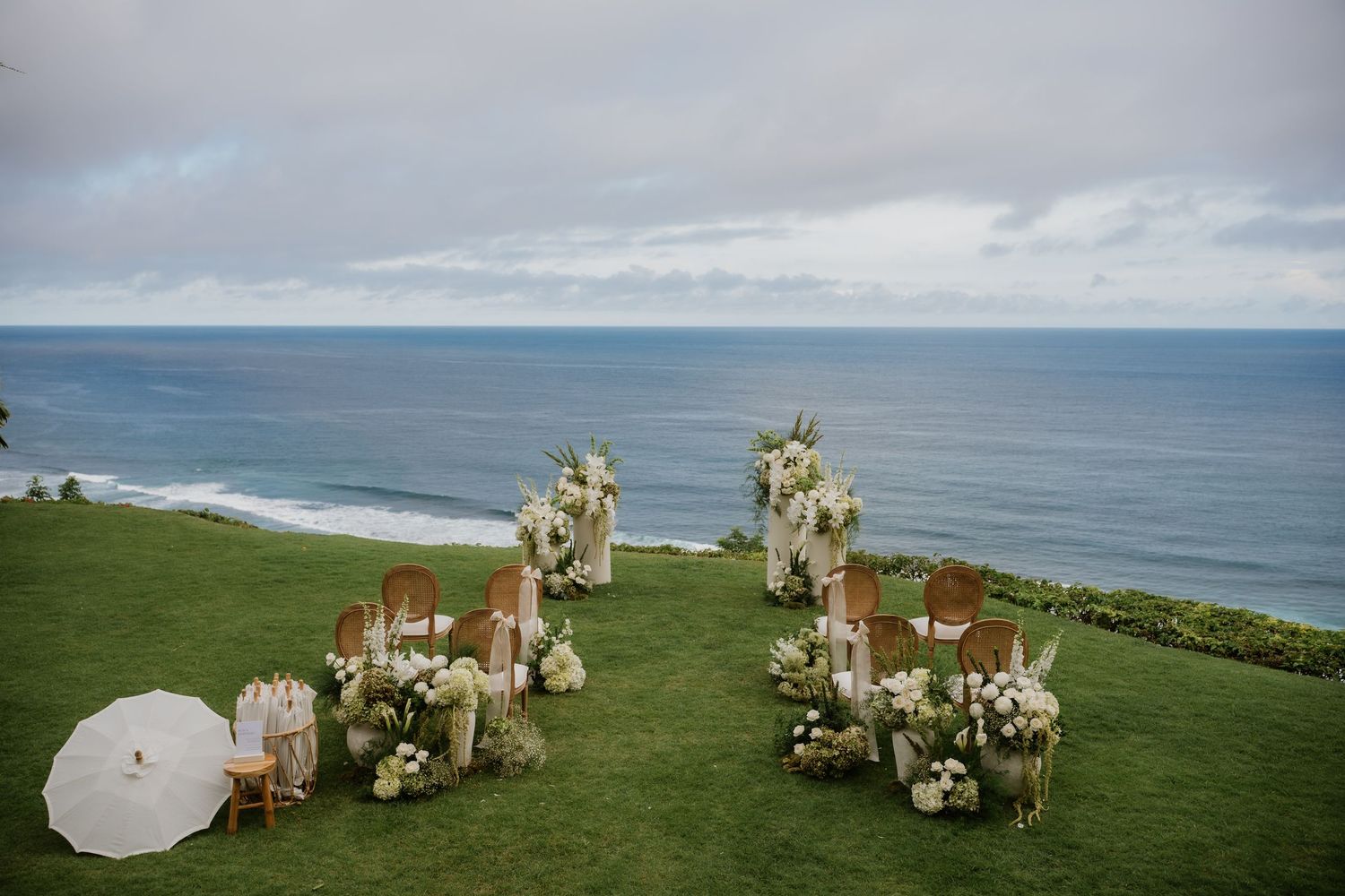 Elegant outdoor wedding ceremony setup on a clifftop lawn overlooking the ocean with white floral arrangements.
