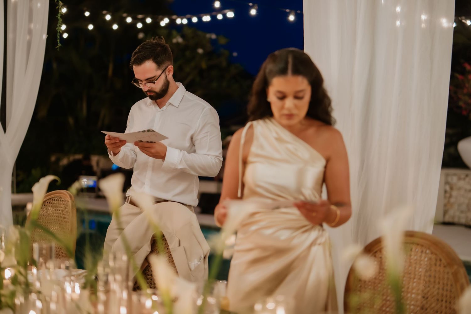 Bride and groom reading vows from cards at elegant candlelit wedding reception with string lights.